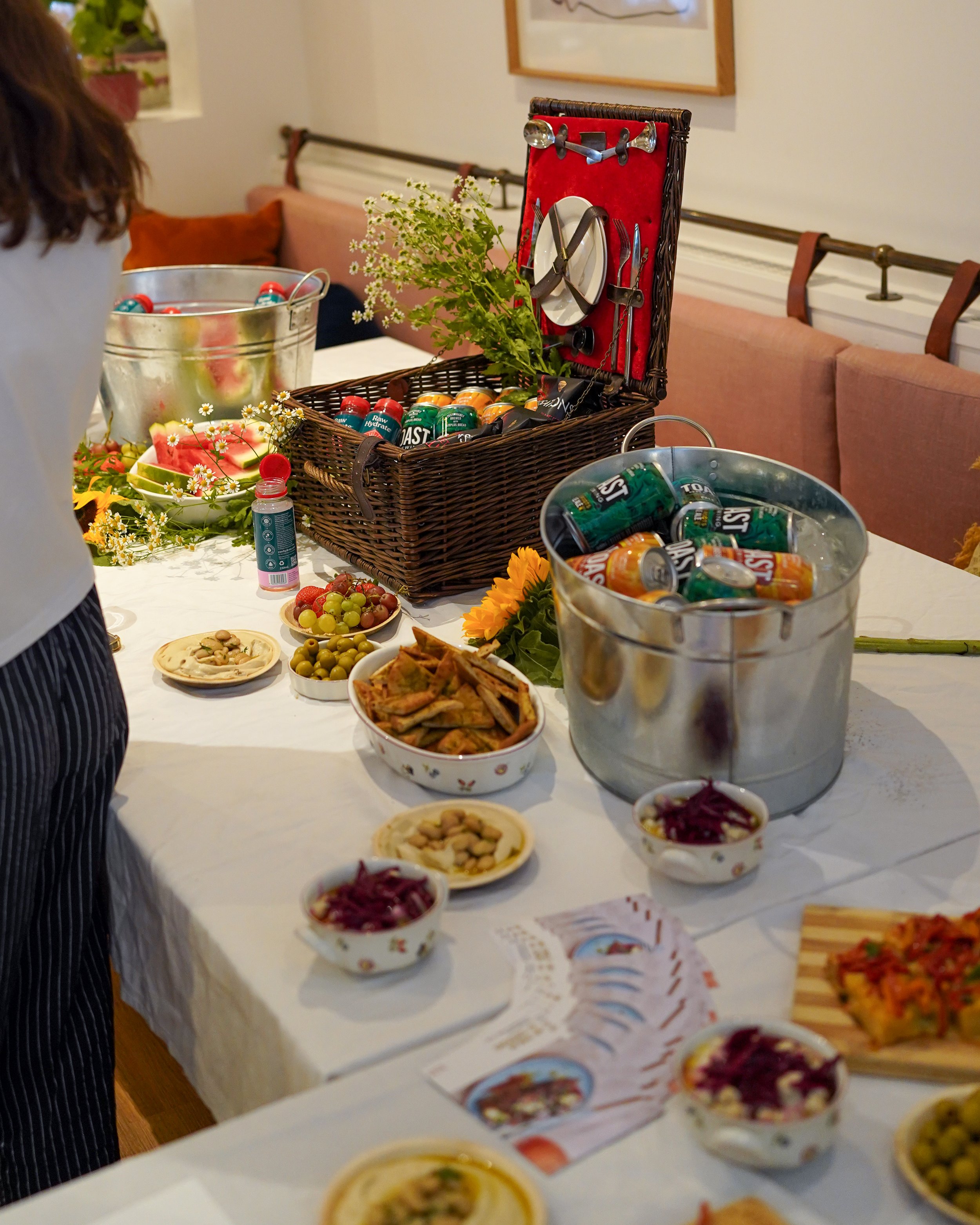 A table with various snacks, dips, and small bowls of food, with a basket containing canned drinks and condiments, set in a cozy indoor setting.