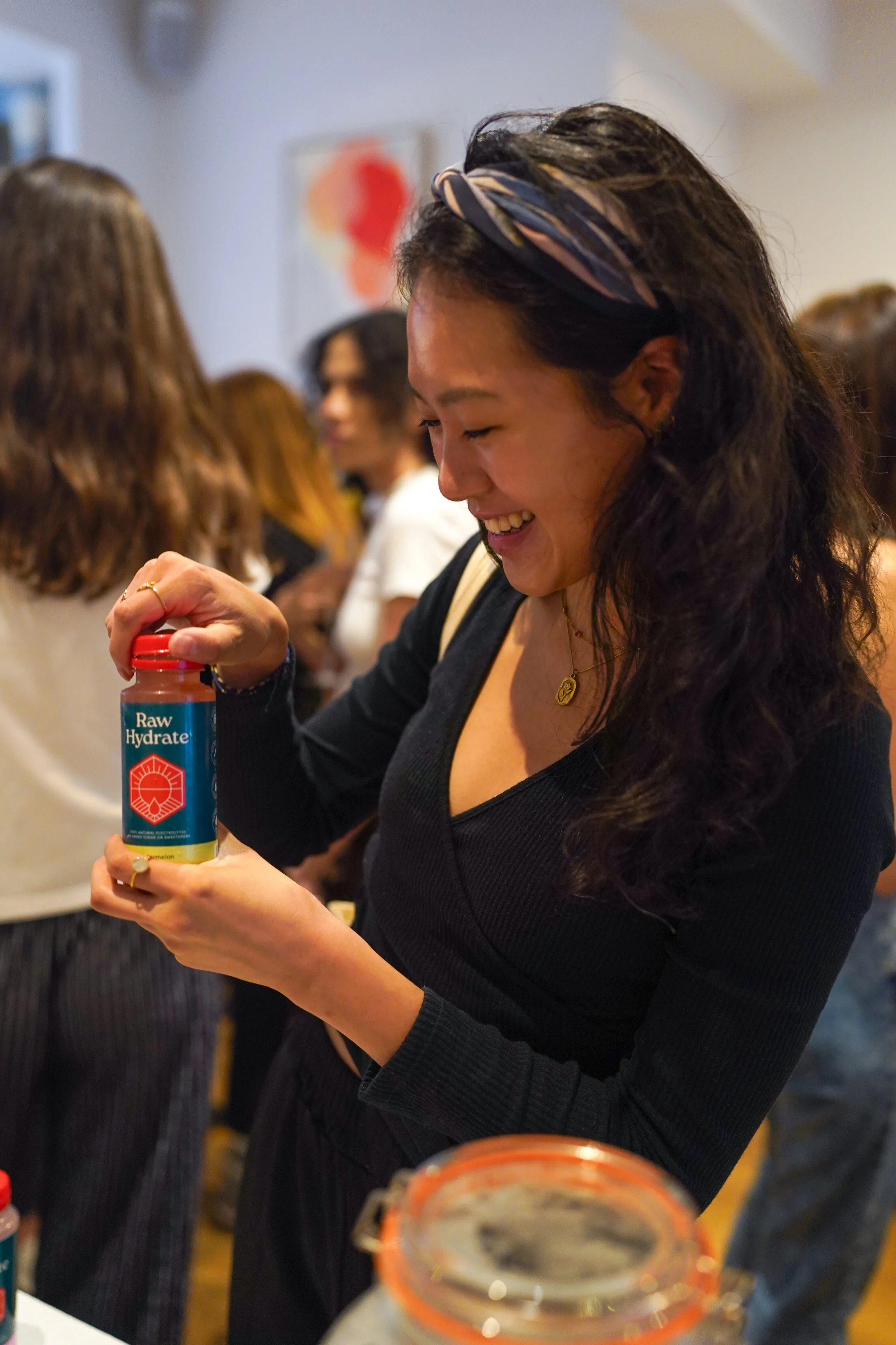 A smiling woman with dark, curly hair and a patterned headband opening a bottle of water at a crowded indoor event.