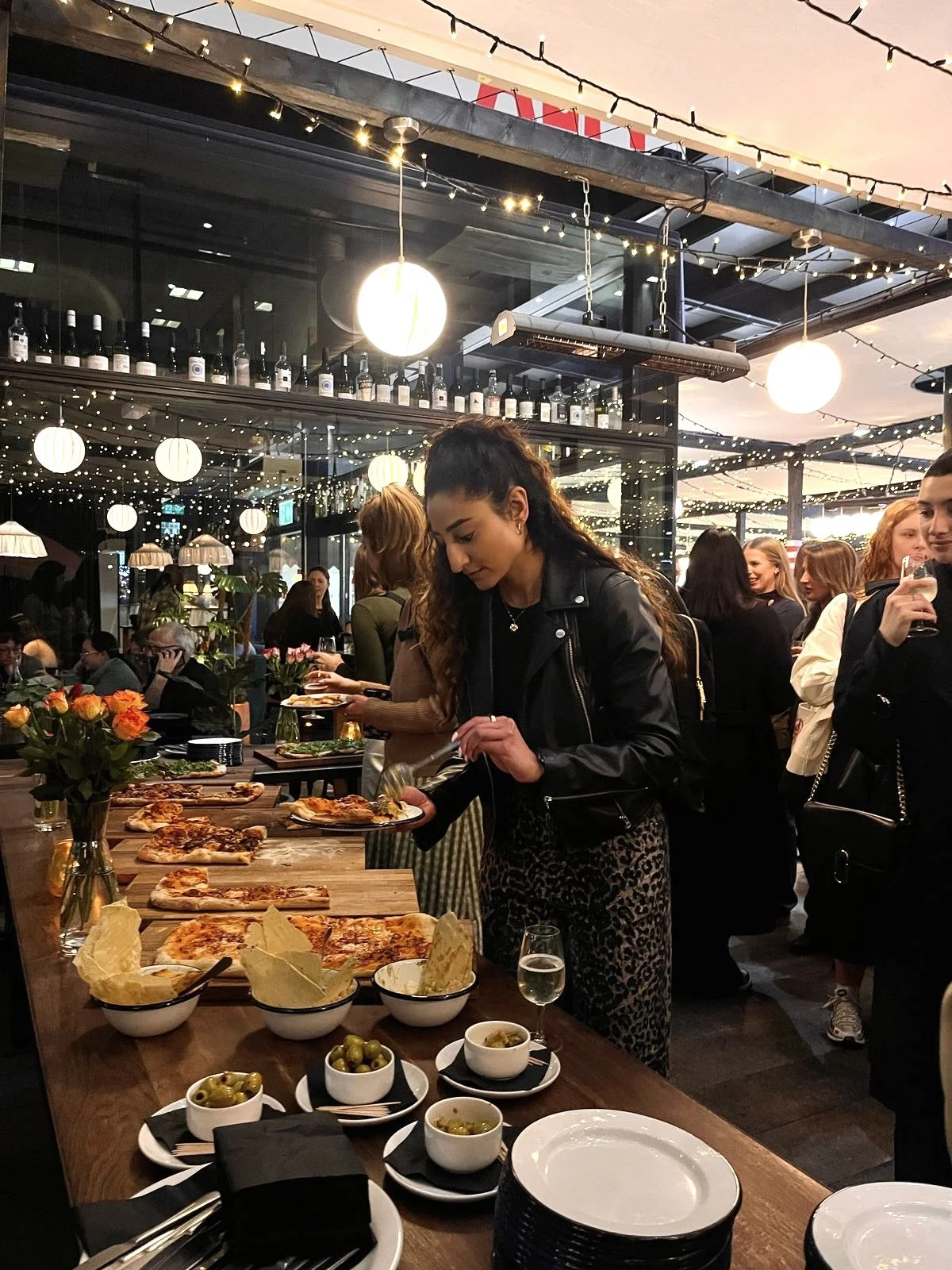 People enjoying a social gathering at a restaurant or bar, with a woman serving pizza and bowls of olives on a long wooden table, decorated with flowers, under warm string lights during sunset.