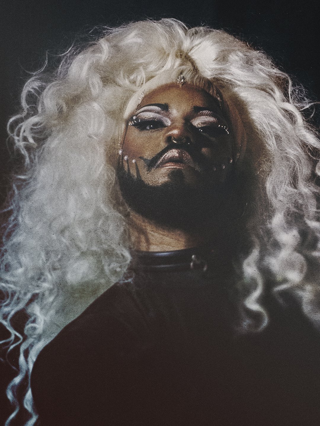 Person with curly white wig and dramatic makeup, wearing black clothing.