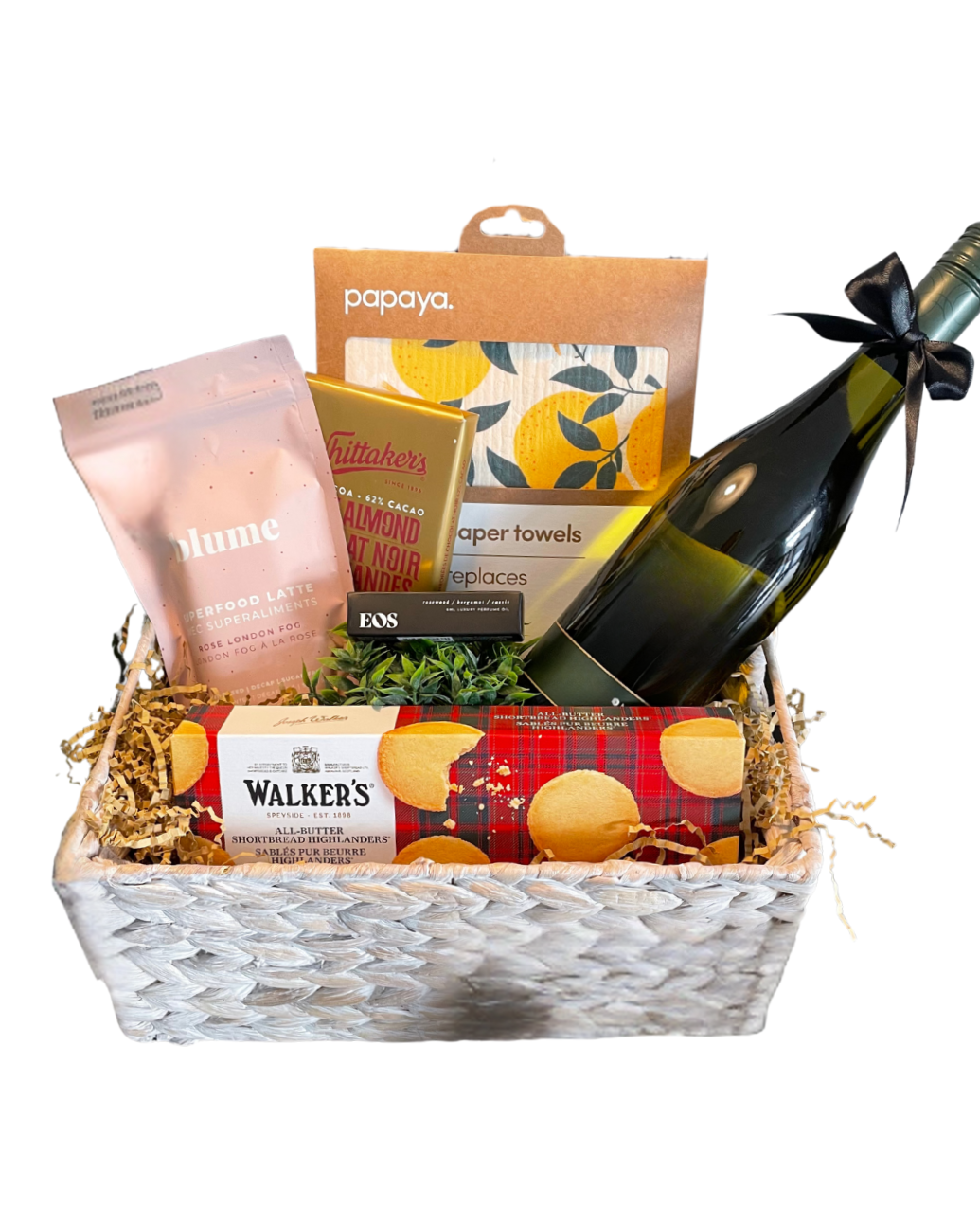 Gift basket with snacks, a bottle of sparkling wine (non alcoholic), reusable paper towels with lemons on them, and a small potted plant, essential oil perfume rollerball, superfood late mix, all arranged in a woven container.