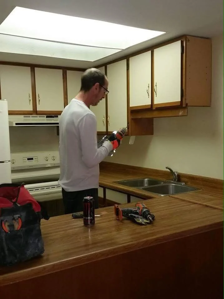 A man working on kitchen cabinets, holding a power drill, in a kitchen with wooden cabinets and a double sink.