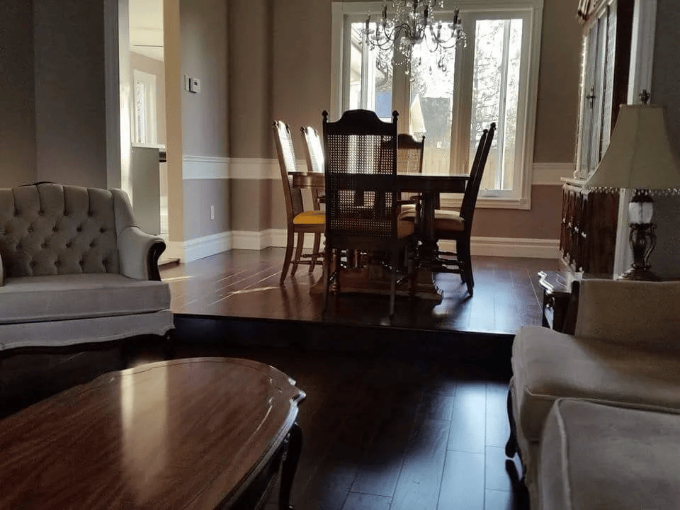 Living room with wooden furniture, including a dining table with six chairs, two armchairs, a coffee table, and a side table with a lamp. Large windows allow natural light into the room.