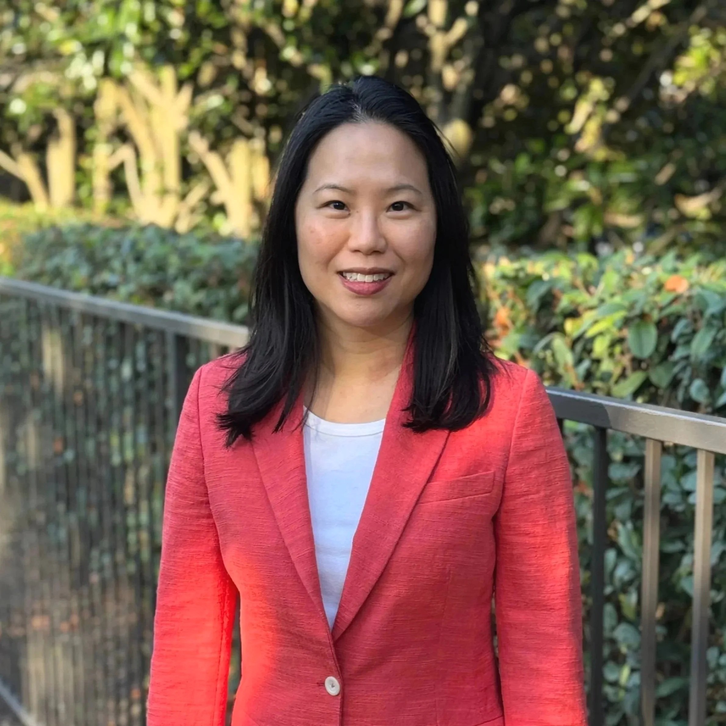 Portrait of a woman with long black hair wearing a coral blazer and a white shirt, standing outdoors in front of green bushes and trees.
