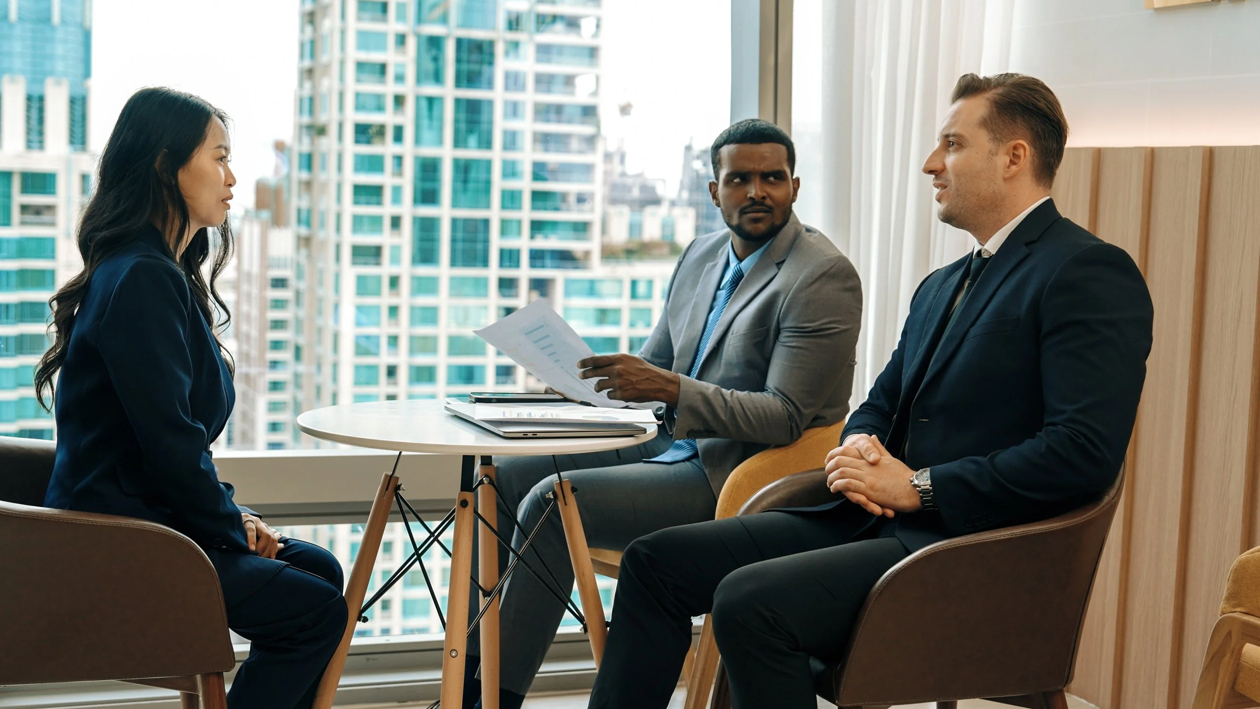 A woman and two men engaged in a conversation in a modern office with large windows and a city view, seated on chairs around a small round table with documents.