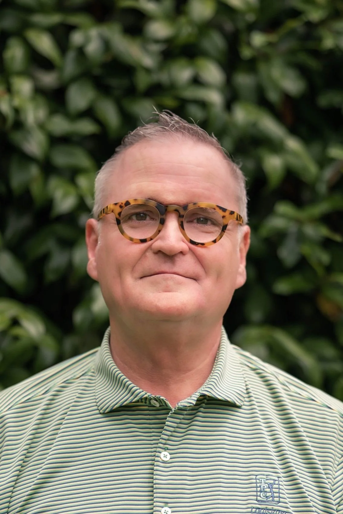 A man wearing tortoise shell glasses and a green striped collared shirt stands in front of green foliage.