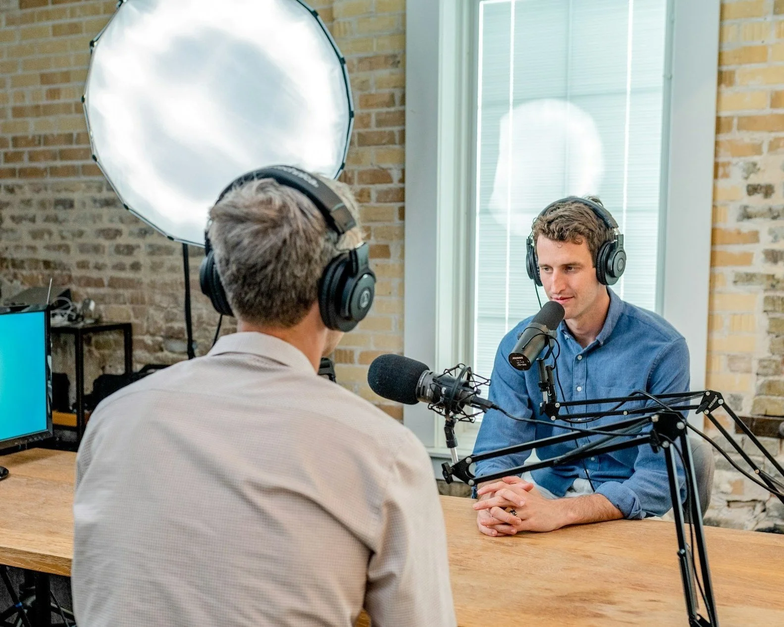 Two men wearing headphones are having a podcast in a studio with brick walls, a large softbox light, and a wooden table; one man is speaking into a microphone while the other listens.