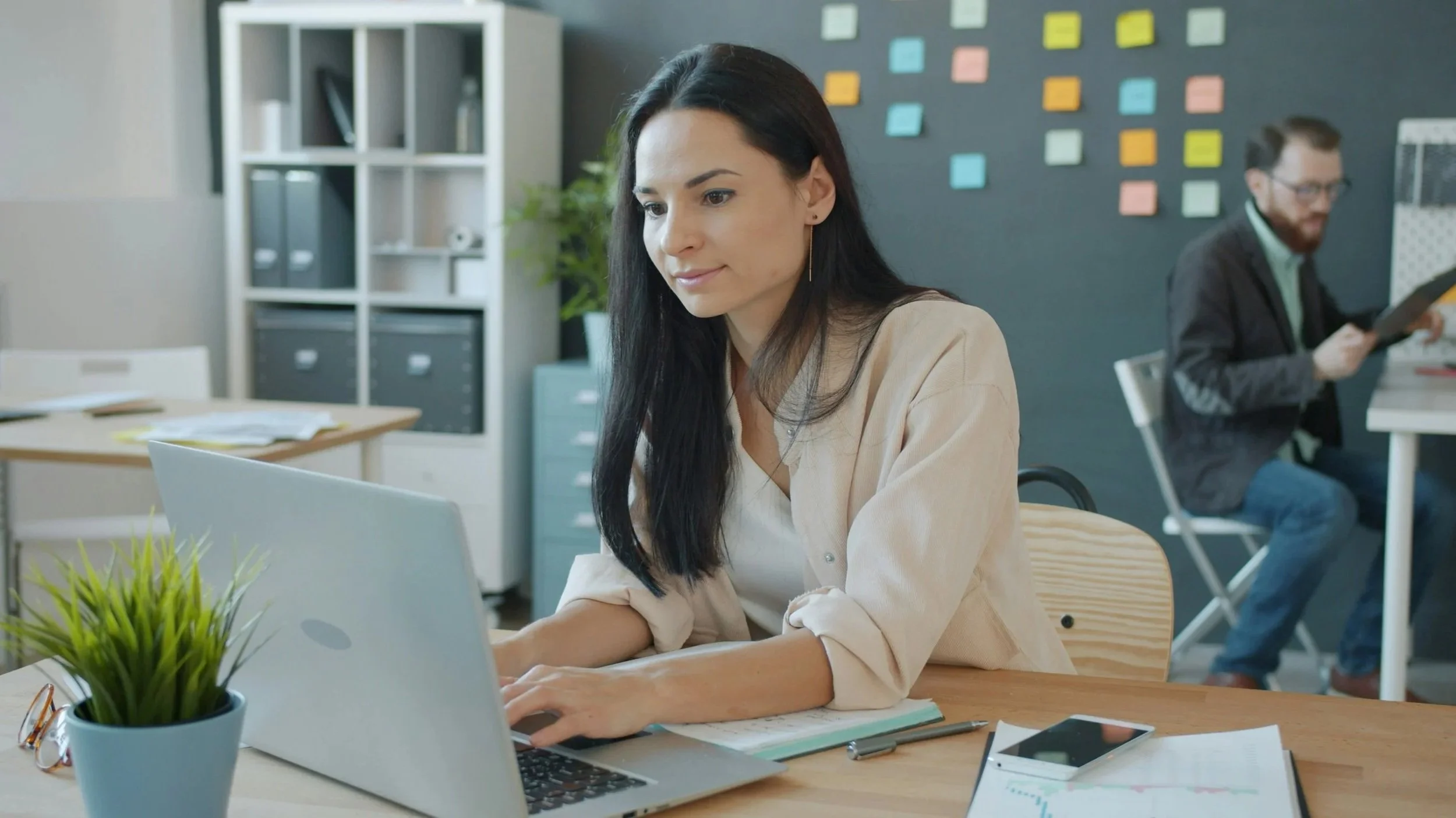 A woman working on a laptop at a desk in an office with a man in the background reading a document