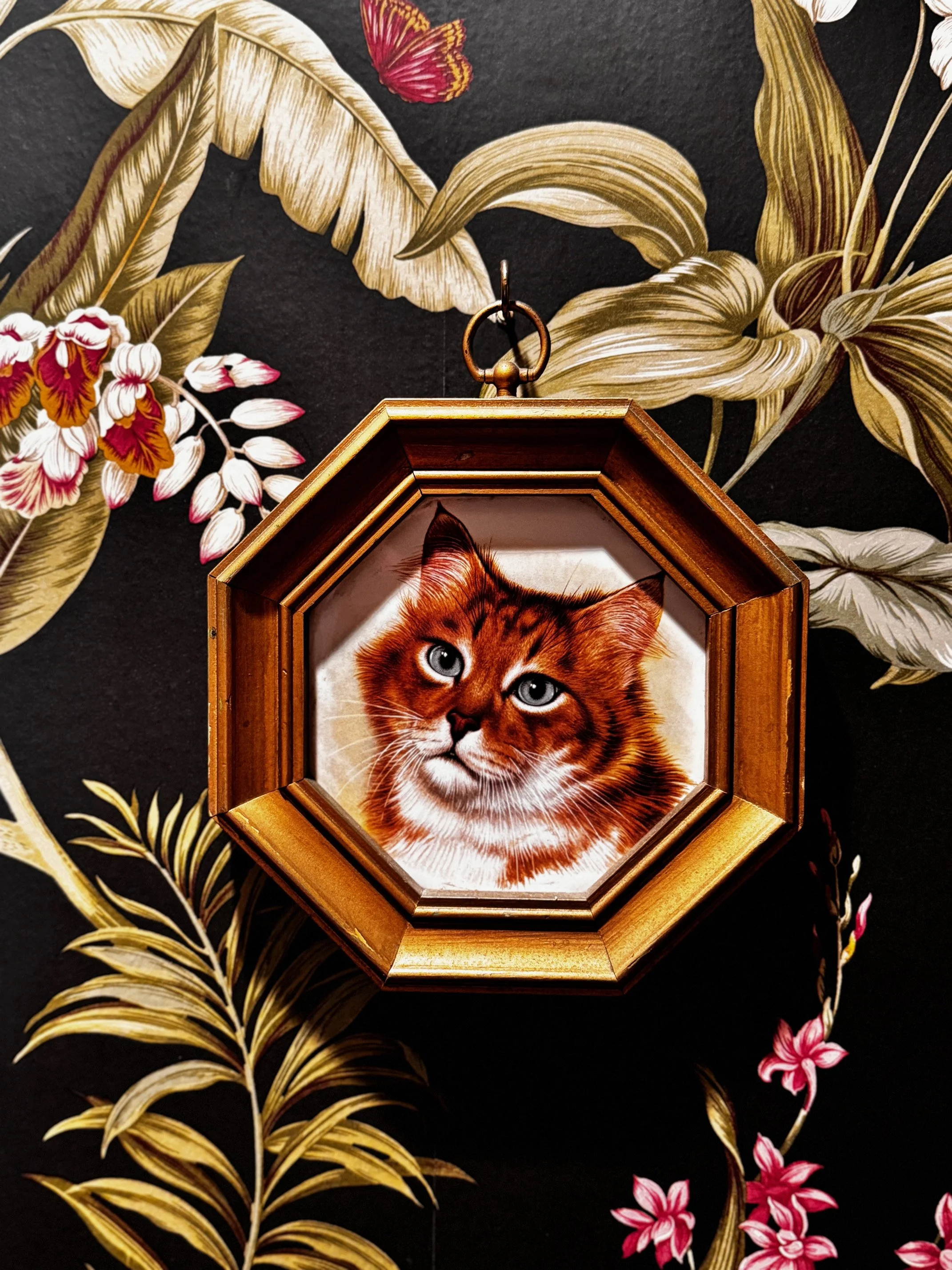 Vintage-style wall art depicting a framed painting of a cat against floral wallpaper background.