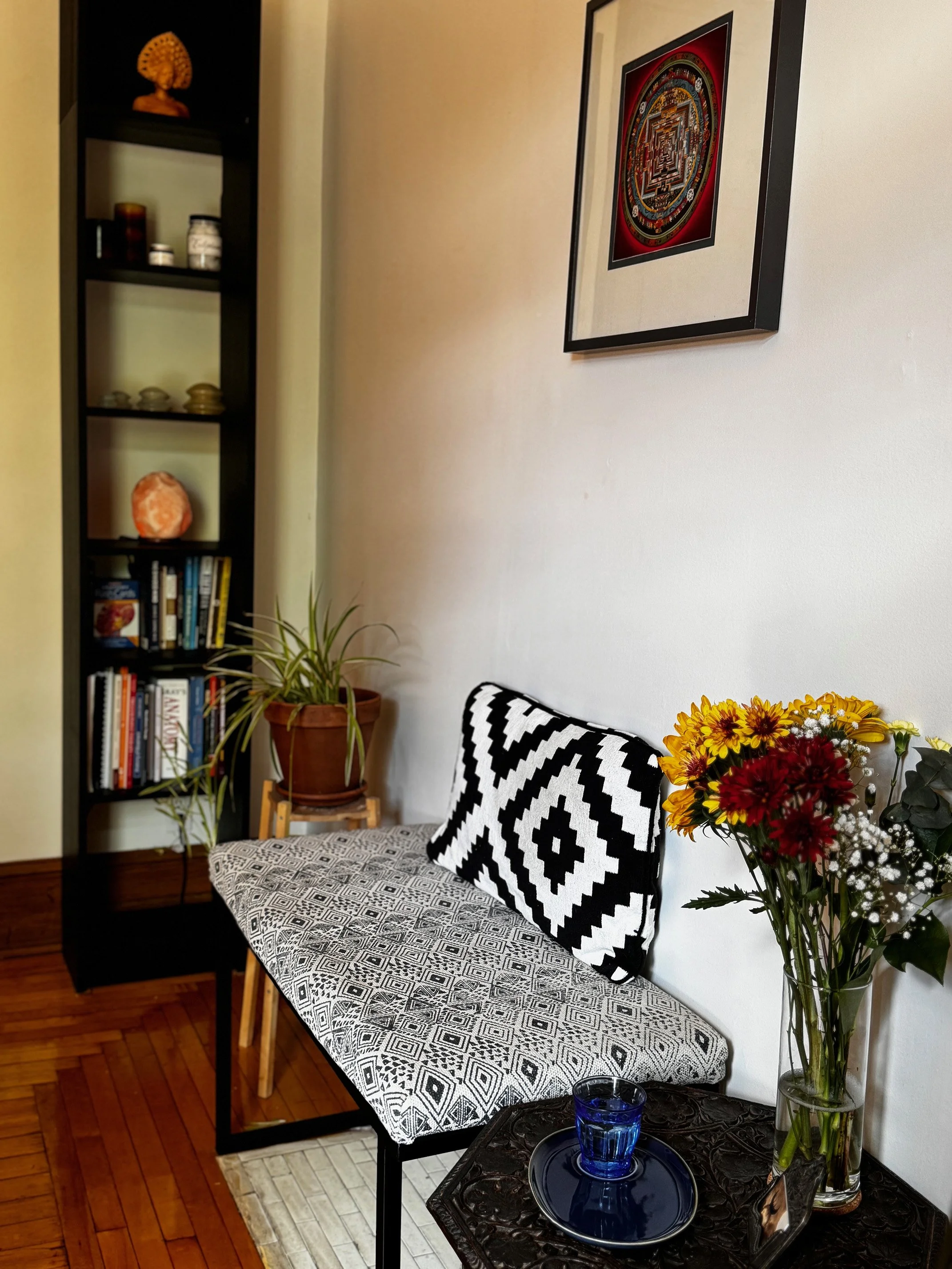 Welcoming seat at Mile End Massage where clients discuss their treatment and prepare for their session. The space features a patterned bench with a black and white cushion, wall art, a bookshelf filled with books and decor including a glowing salt la