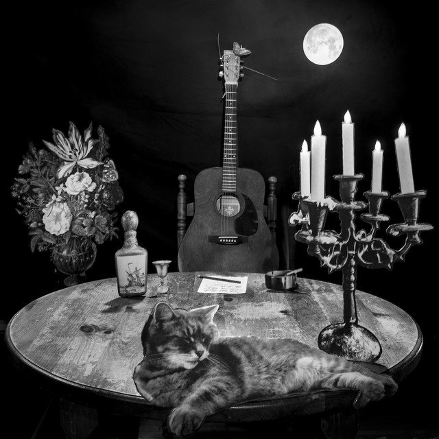 Album cover - Songs from the Round Table. Black and white image of a cat relaxing on a round wooden table with a guitar in the background, a floral arrangement, a candlestick with lit candles, and a full moon visible above.
