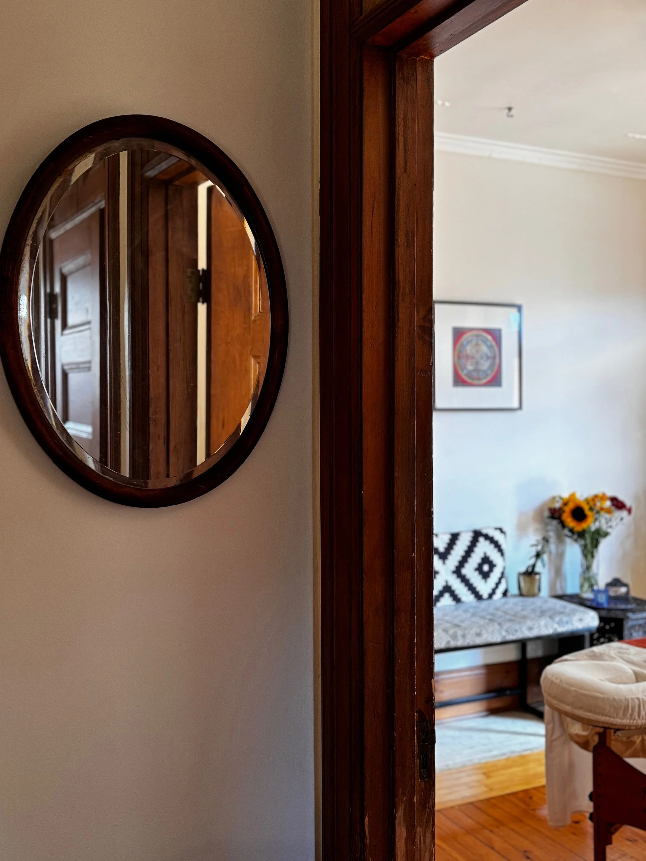 Entrance view into the massage room at Mile End Massage, featuring an elegant oval mirror by the doorway. Beyond, a glimpse of the welcoming bench seat beside a side table with fresh flowers, setting a serene tone for relaxation.
