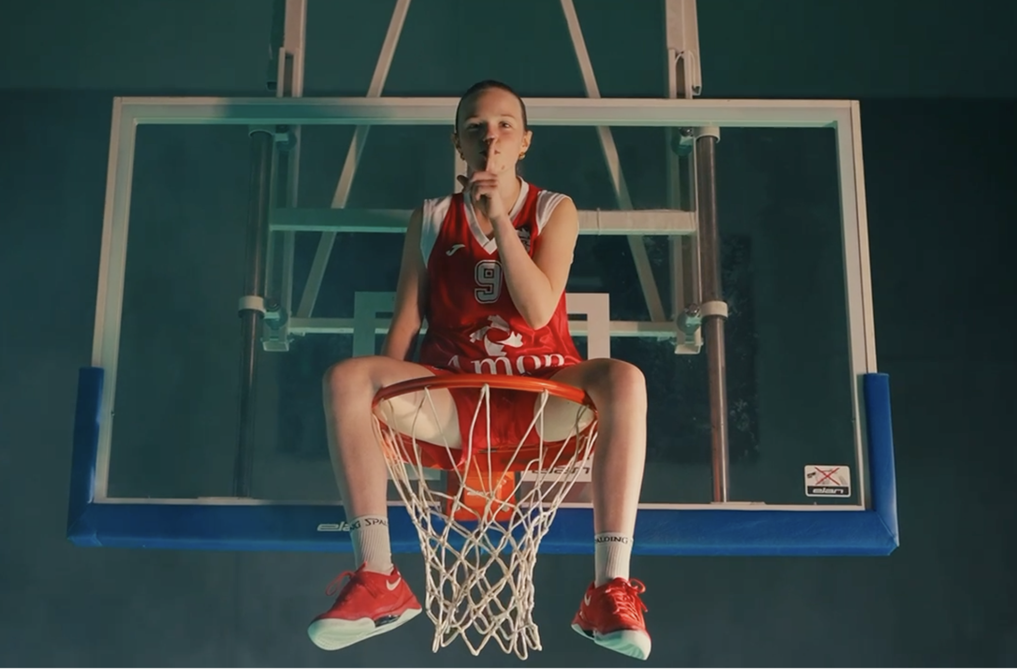 A female basketball player in a red uniform sitting on a basketball hoop stand, holding her finger to her lips in a shushing gesture, with the camera pointing up from below the basket.