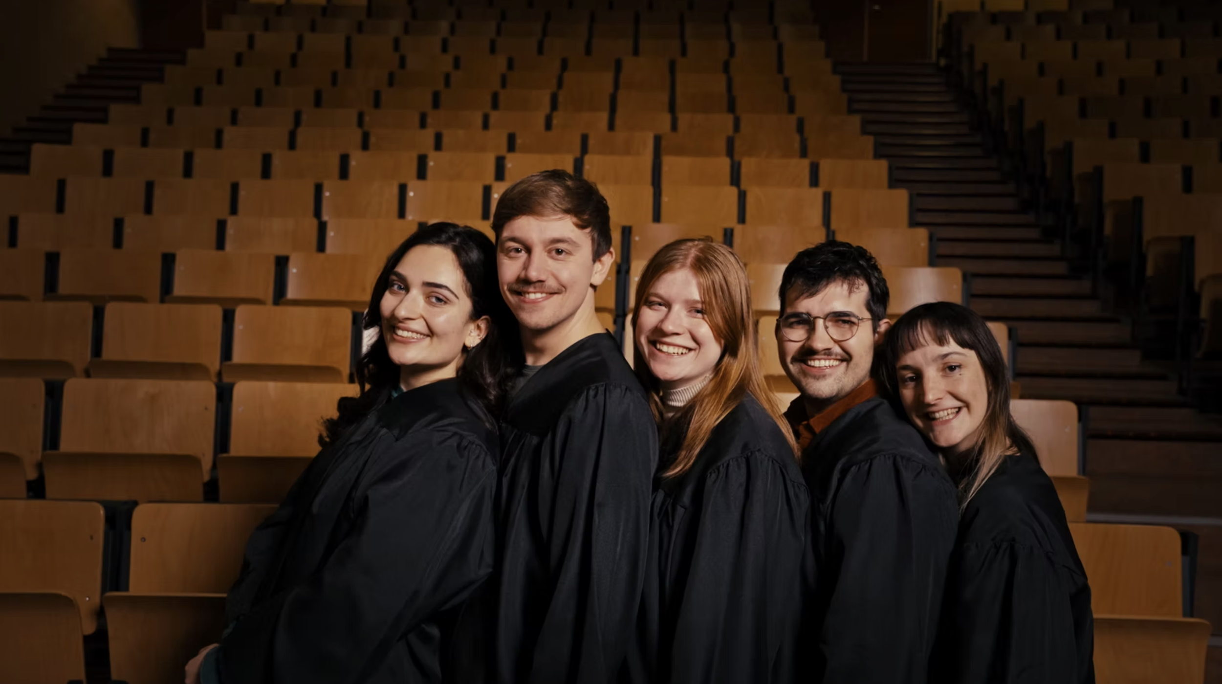 Group of five graduates in black gowns smiling in an auditorium with rows of empty wooden seats behind them.
