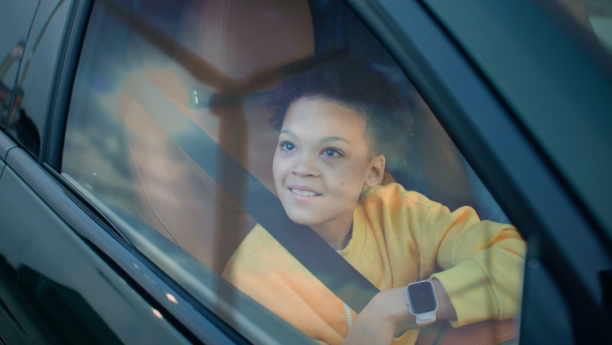A young boy with curly hair and a yellow shirt sitting in the driver's seat of a car, looking out the window with a smile.