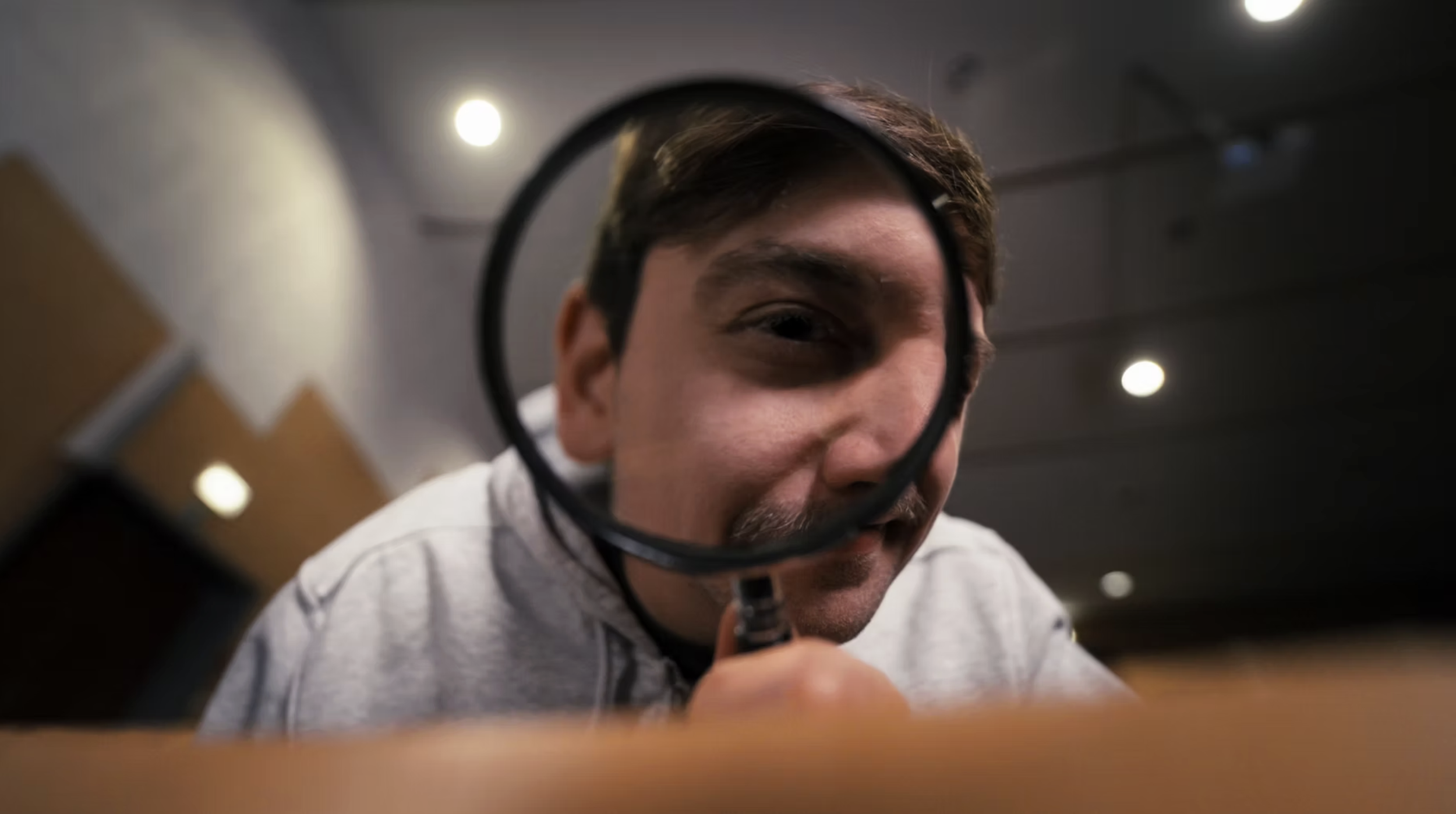 A young man with brown hair and a mustache, holding a magnifying glass in front of his face, looking through it, in an indoor setting with ceiling lights.