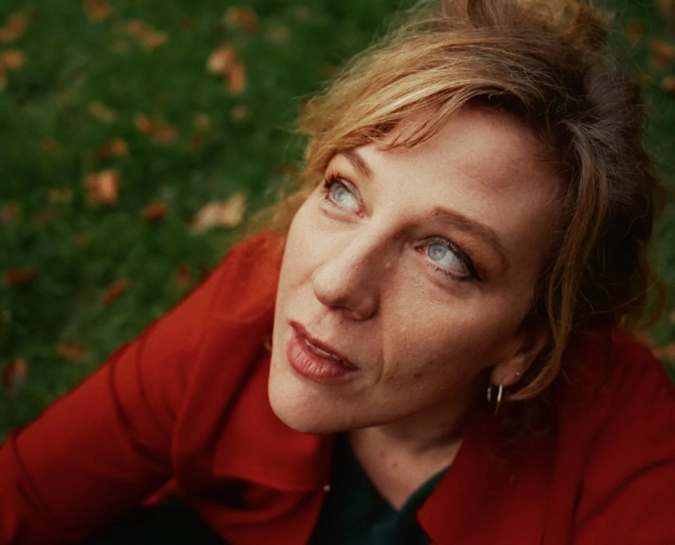 Close-up of a woman with red hair, blue eyes, wearing a red jacket, looking upwards. She has a contemplative expression, with green grass and fallen leaves in the blurred background.
