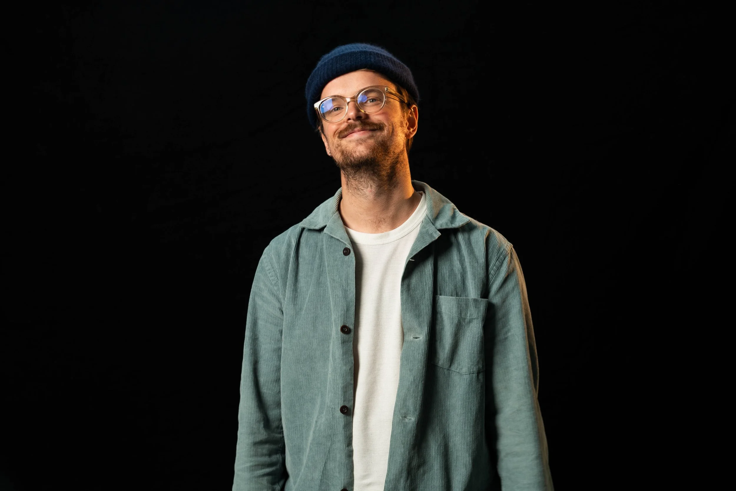 A young man with glasses, a beard, and a mustache wearing a dark beanie, a light-green corduroy jacket, and a white t-shirt, smiling against a black background.