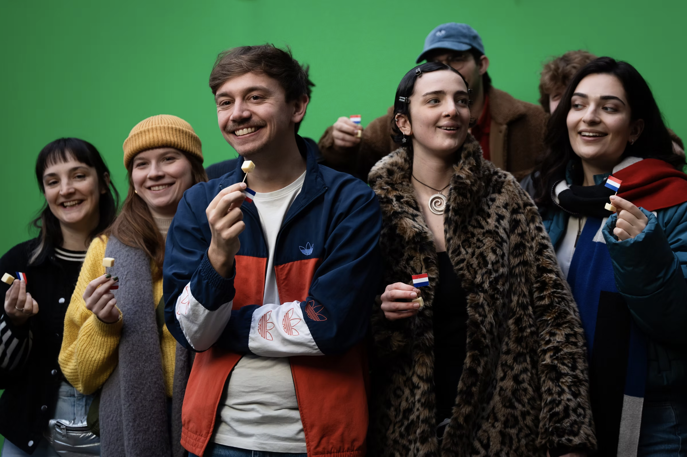 A group of diverse young people smiling and holding Dutch flags and candies in front of a green screen background.