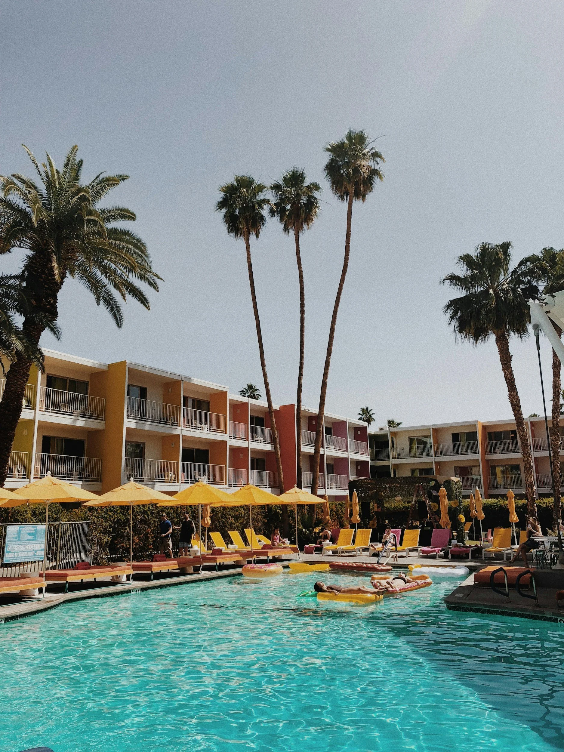 People relaxing and swimming in a hotel swimming pool with yellow umbrellas, surrounded by palm trees and colorful apartment buildings.
