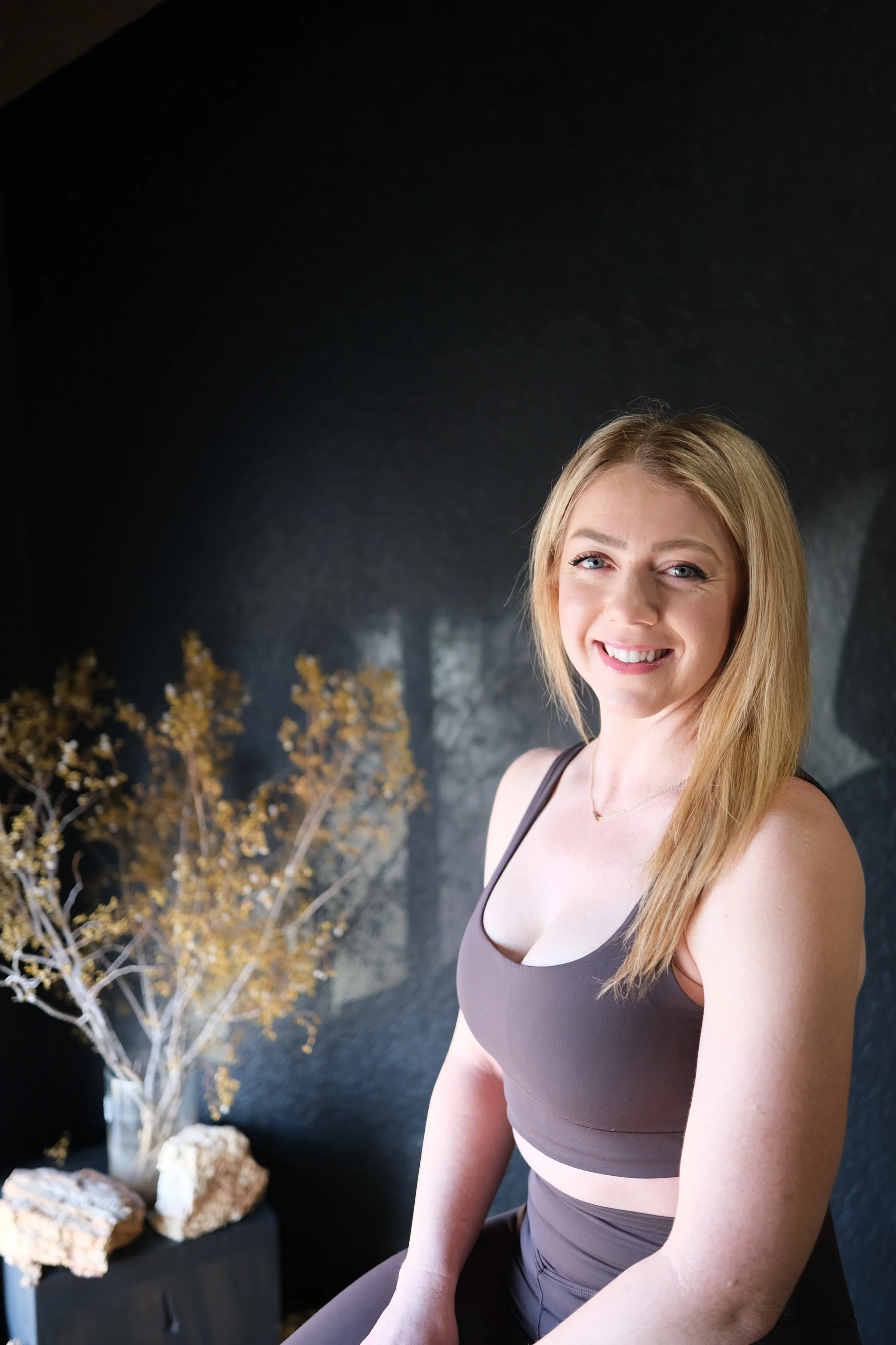A young woman with blonde hair smiling and winking, wearing a black sports bra, sitting indoors with a dark wall, a window, and decorative items including dried plants and rocks in the background.