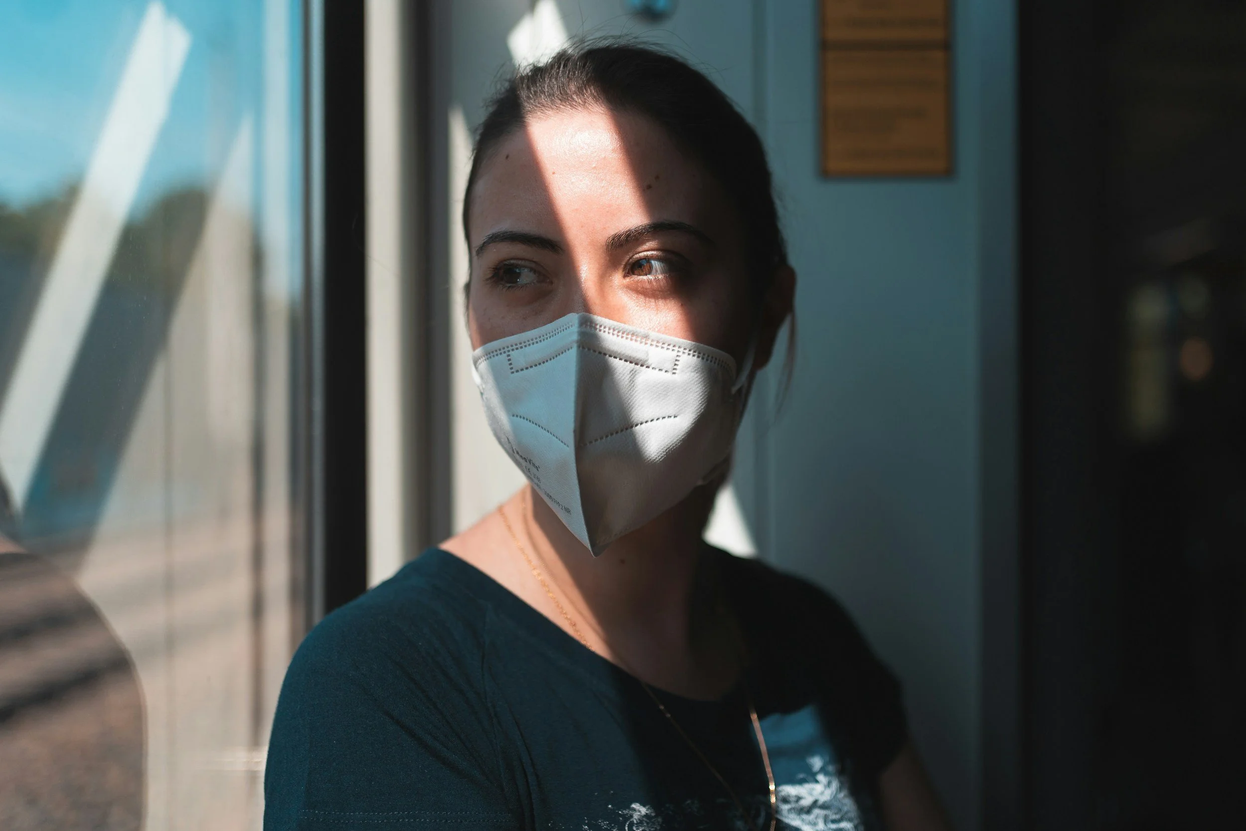 A woman wearing a face mask sitting by a window with sunlight casting a shadow across her face.
