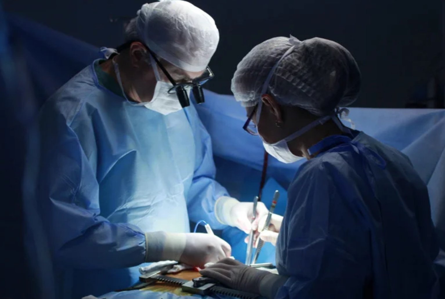 Two surgeons performing an operation in an operating room, wearing surgical gowns, masks, caps, and gloves, focused on the procedure.