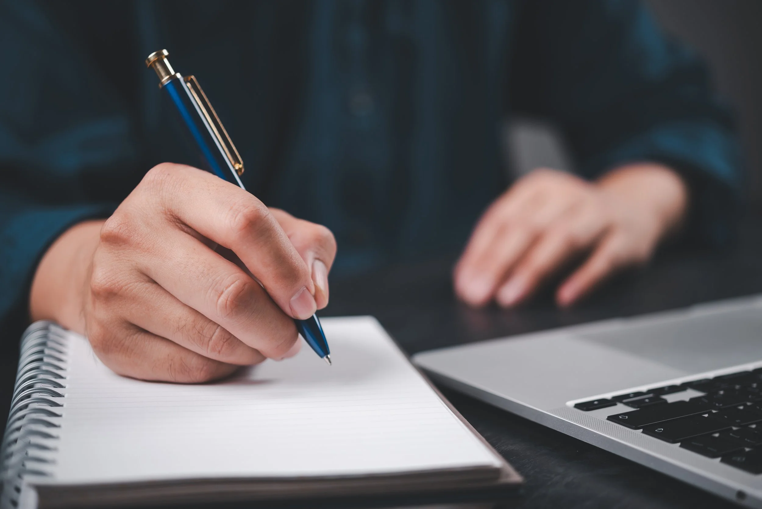 Person writing in a spiral notebook with a blue and gold pen, next to a laptop on a dark desk.