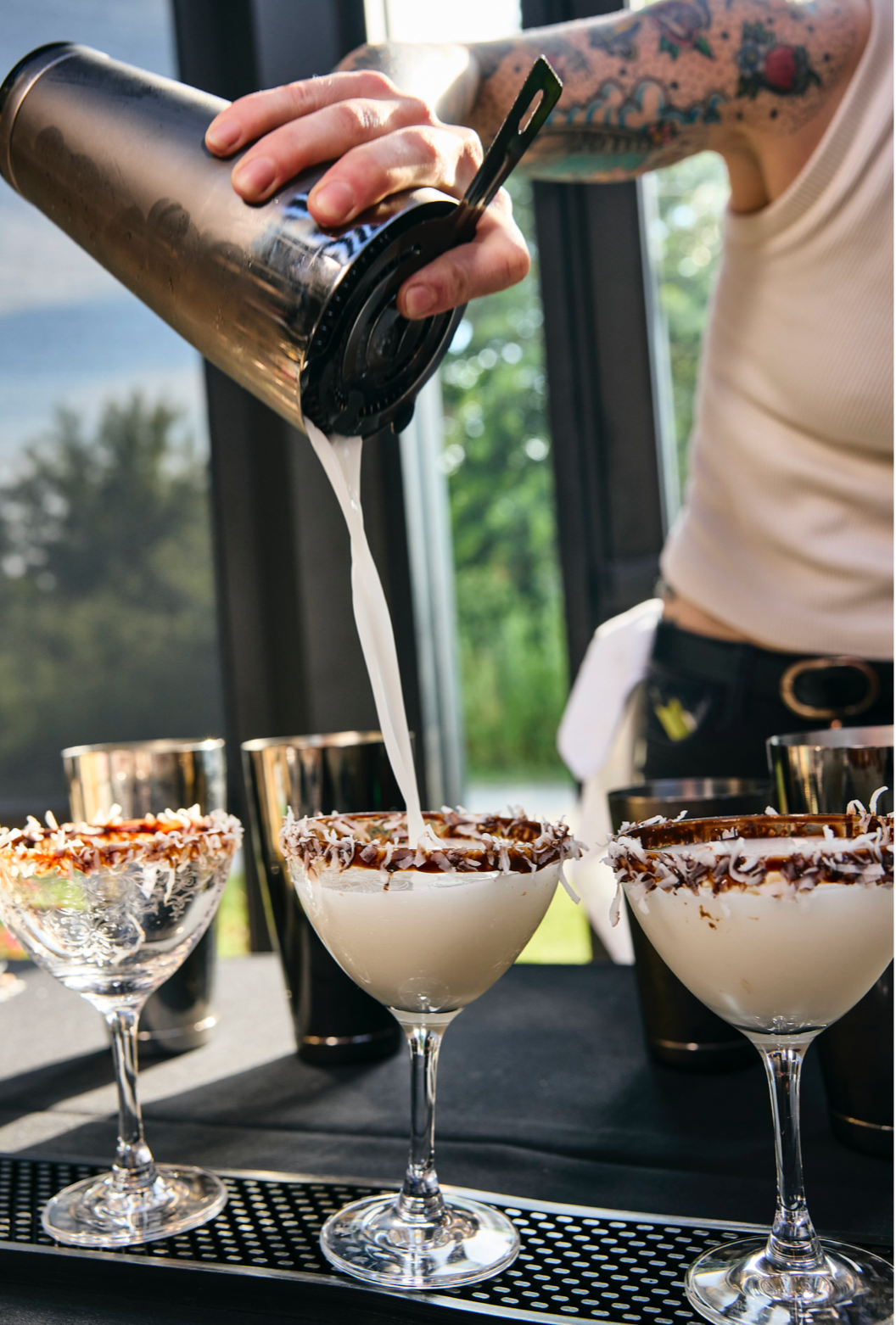 A craft cocktail served in a stemmed glass from Blind Owl Brewery, filled with a pale yellow drink over ice and garnished with a lime wedge. The glass rests on a wooden board surrounded by fresh limes and scattered lavender buds.