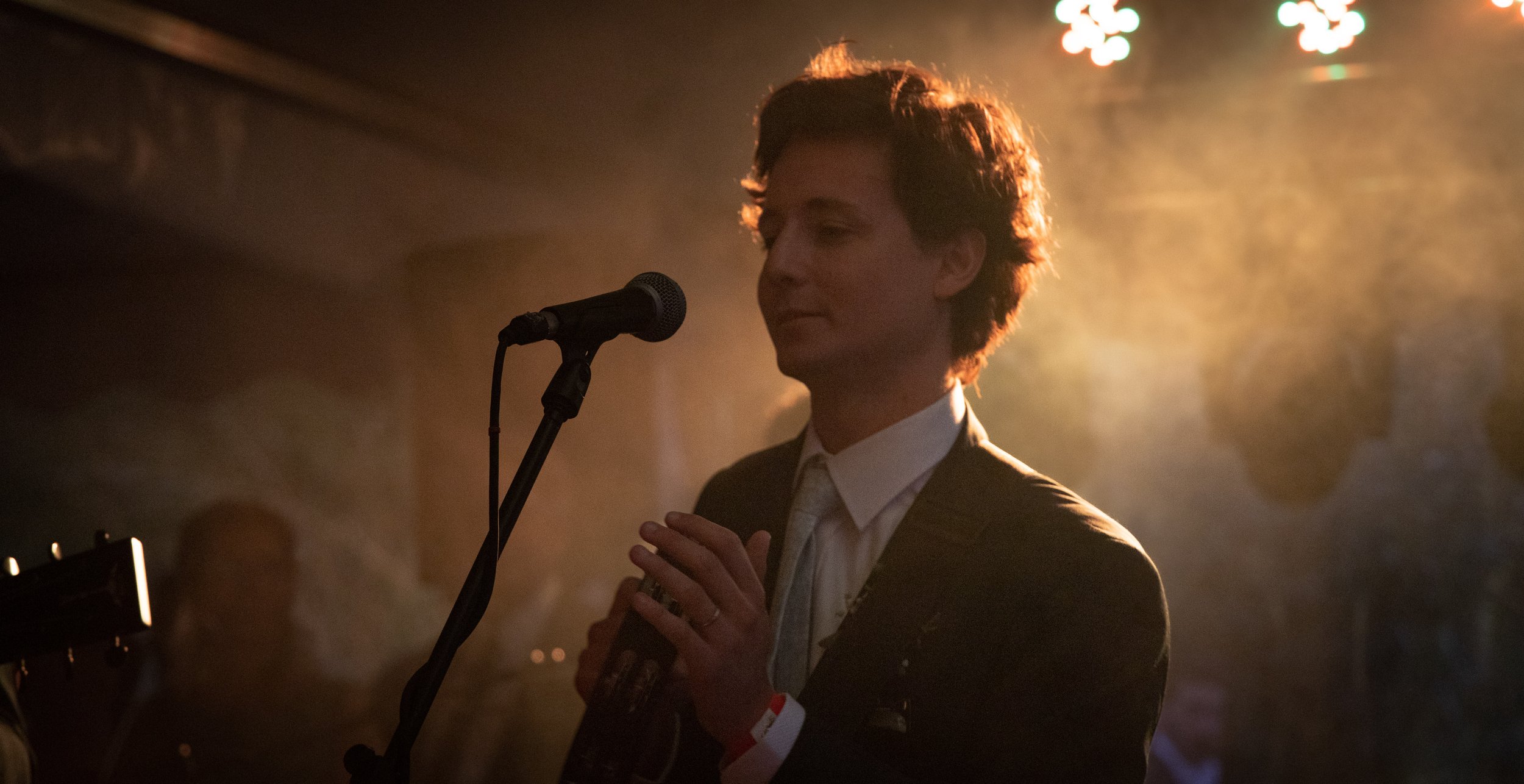 Hombre vestido formalmente sosteniendo una guitarra frente a un micrófono, en un escenario con luces brillantes y ambiente oscuro.