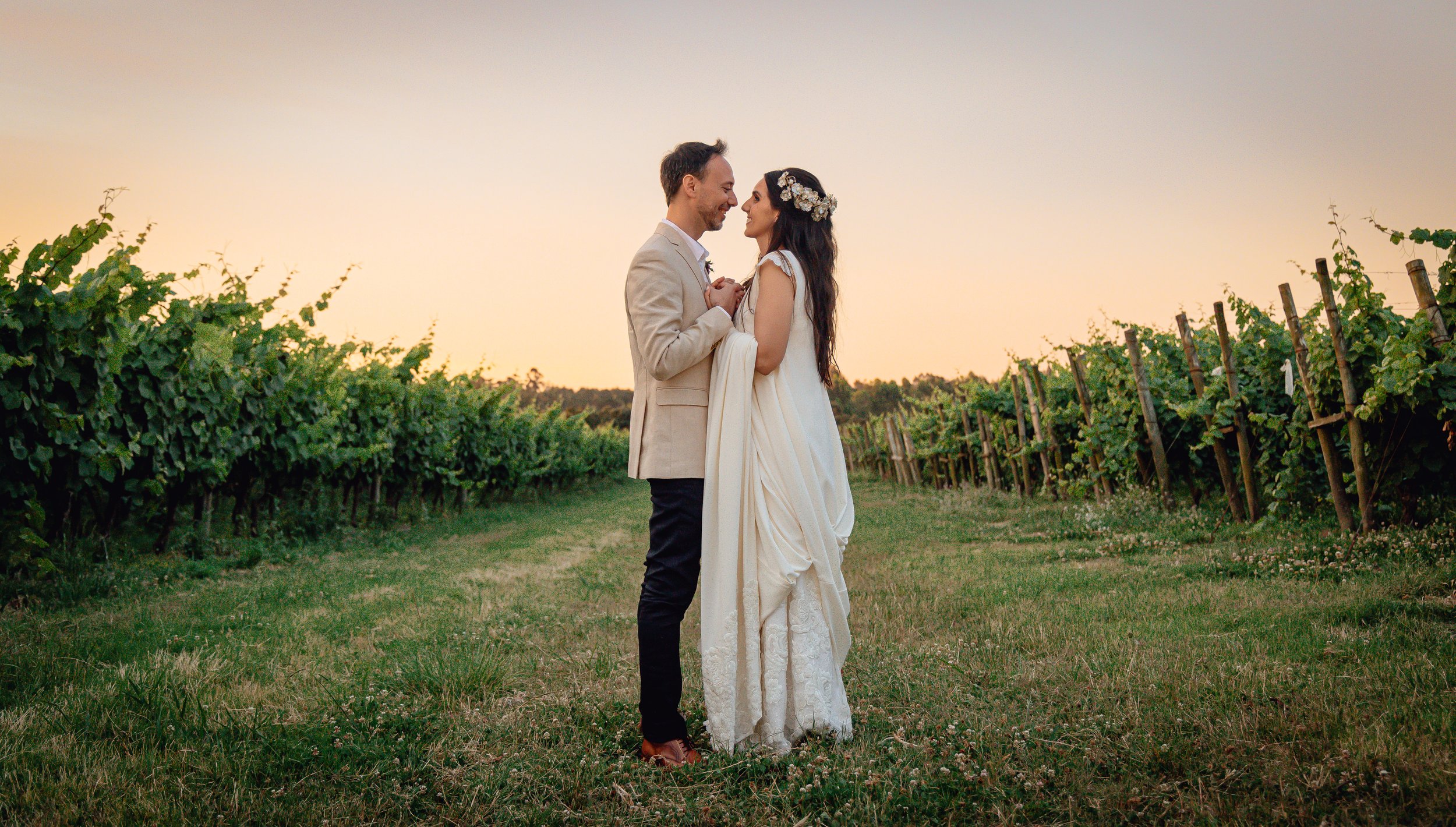 Pareja en un viñedo al atardecer, vestidos elegantemente, mirándose uno al otro con afecto.
