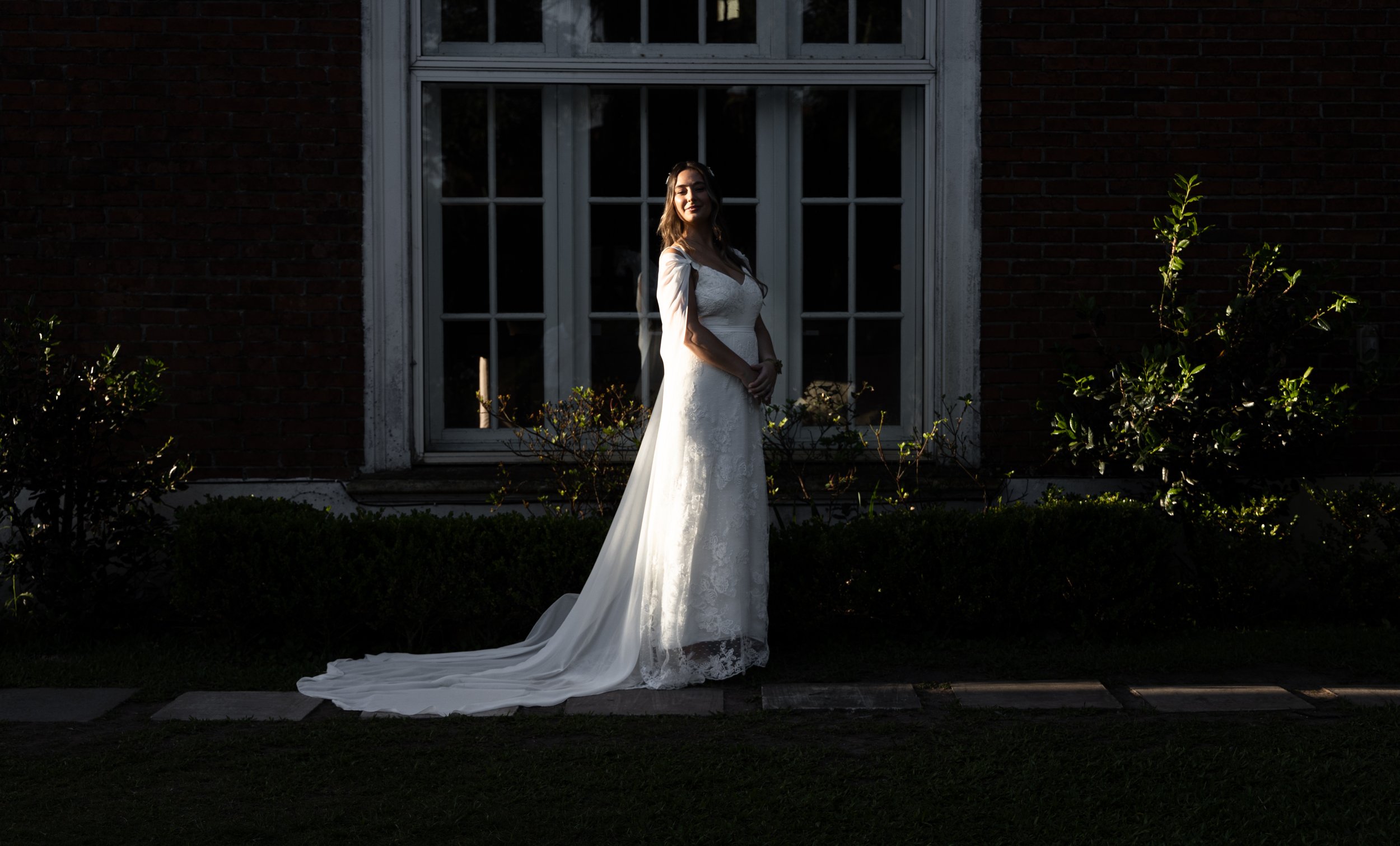 Mujer con vestido de novia posando frente a una ventana grande al aire libre.