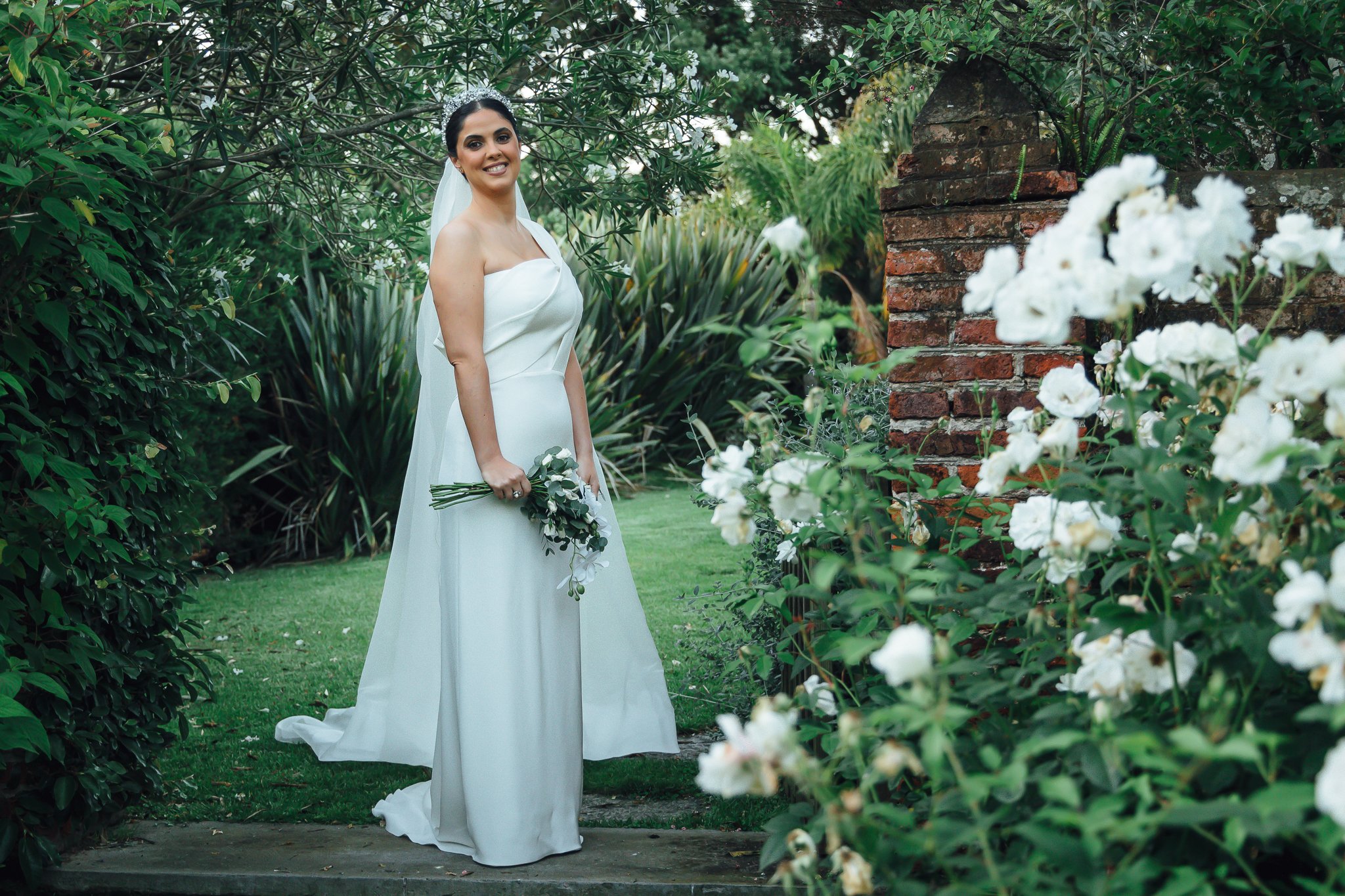 Novia con vestido blanco y ramo de flores, de pie en un jardín con plantas y flores blancas, cerca de un muro de ladrillo.