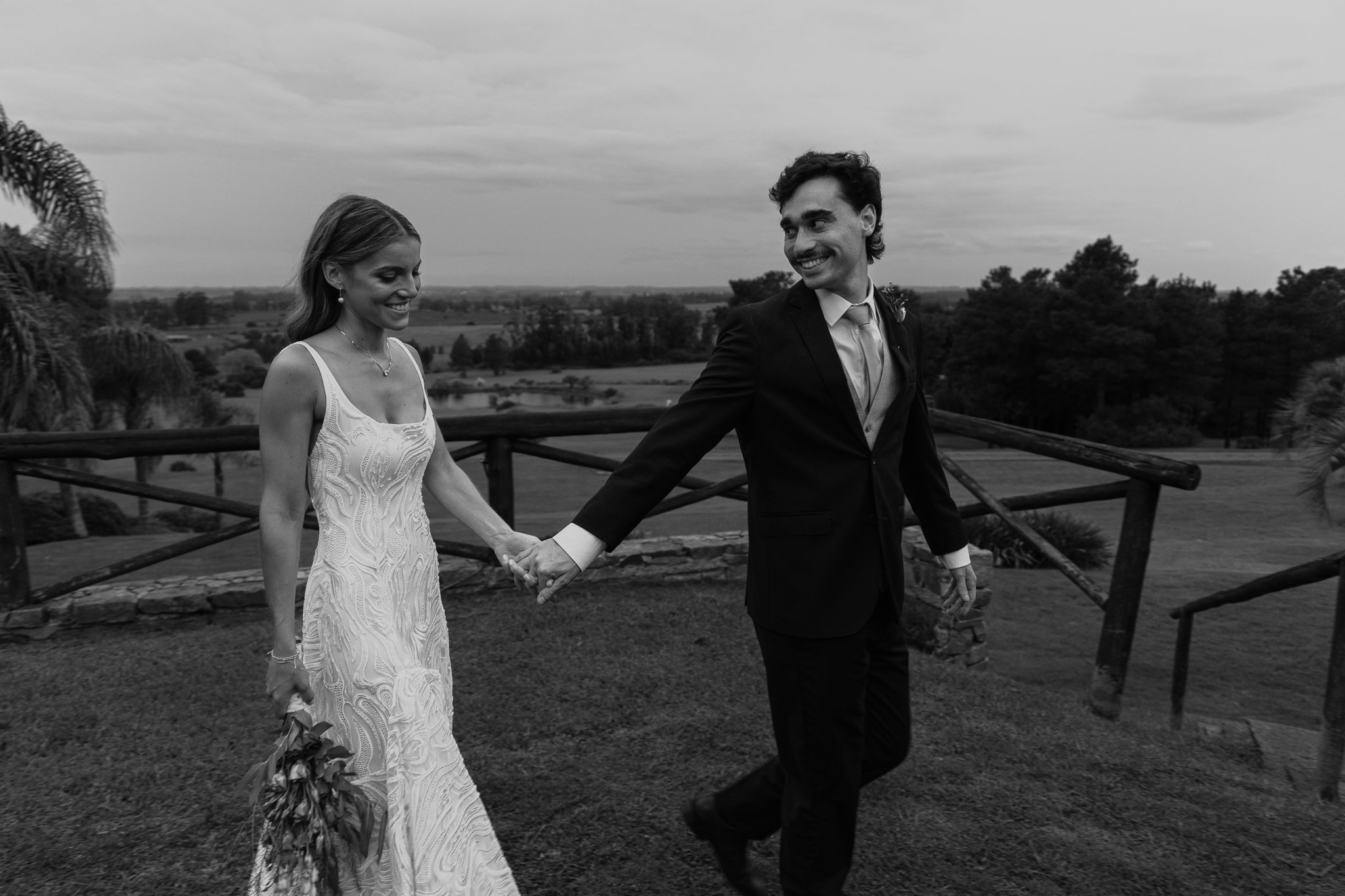Pareja vestida elegantemente caminando de la mano en un entorno natural, la mujer lleva un vestido blanco largo y el hombre un traje oscuro. Escena en blanco y negro.