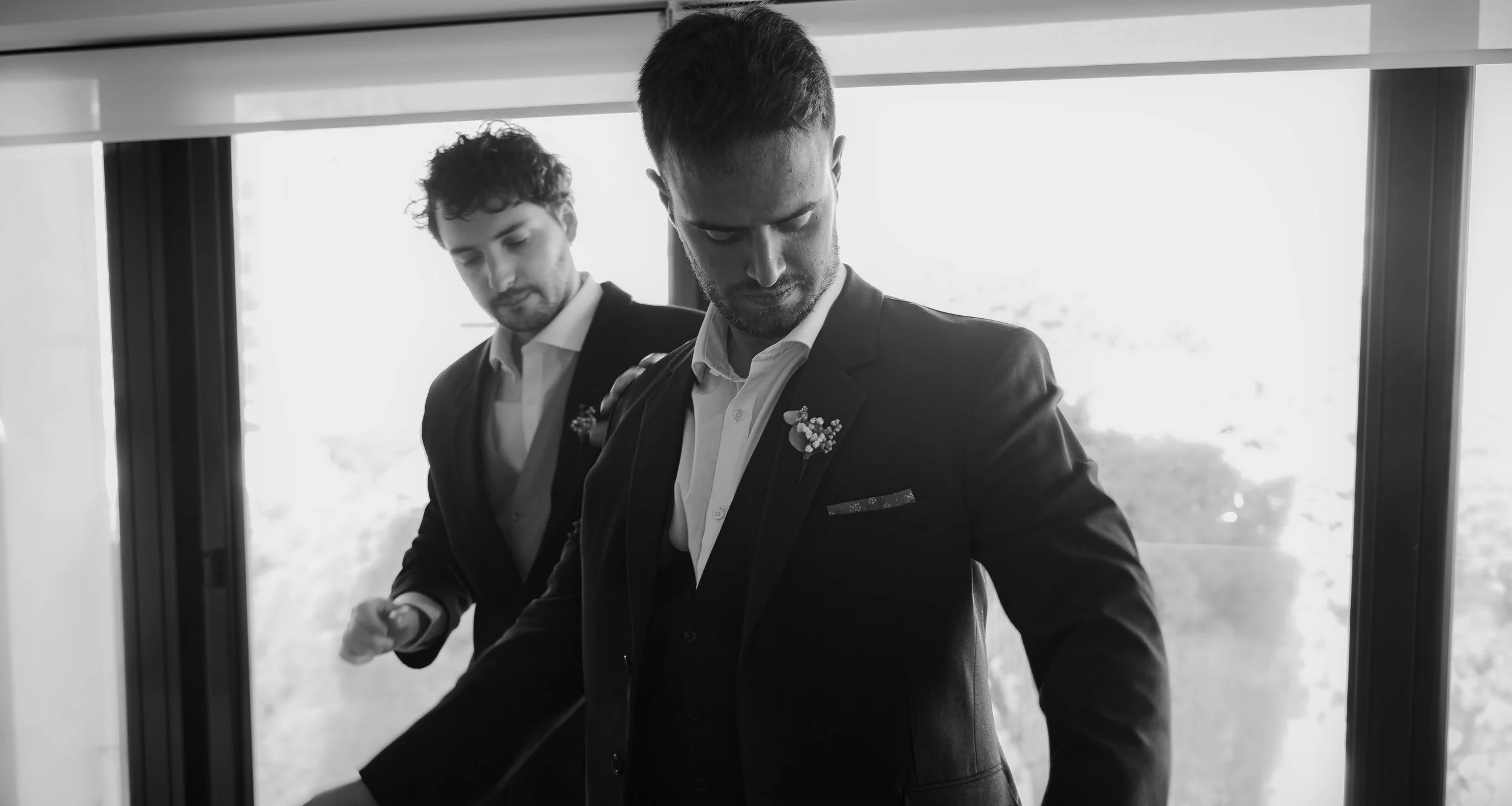 Dos hombres vistiendo trajes formales, posiblemente preparándose para un evento especial, en un entorno interior.