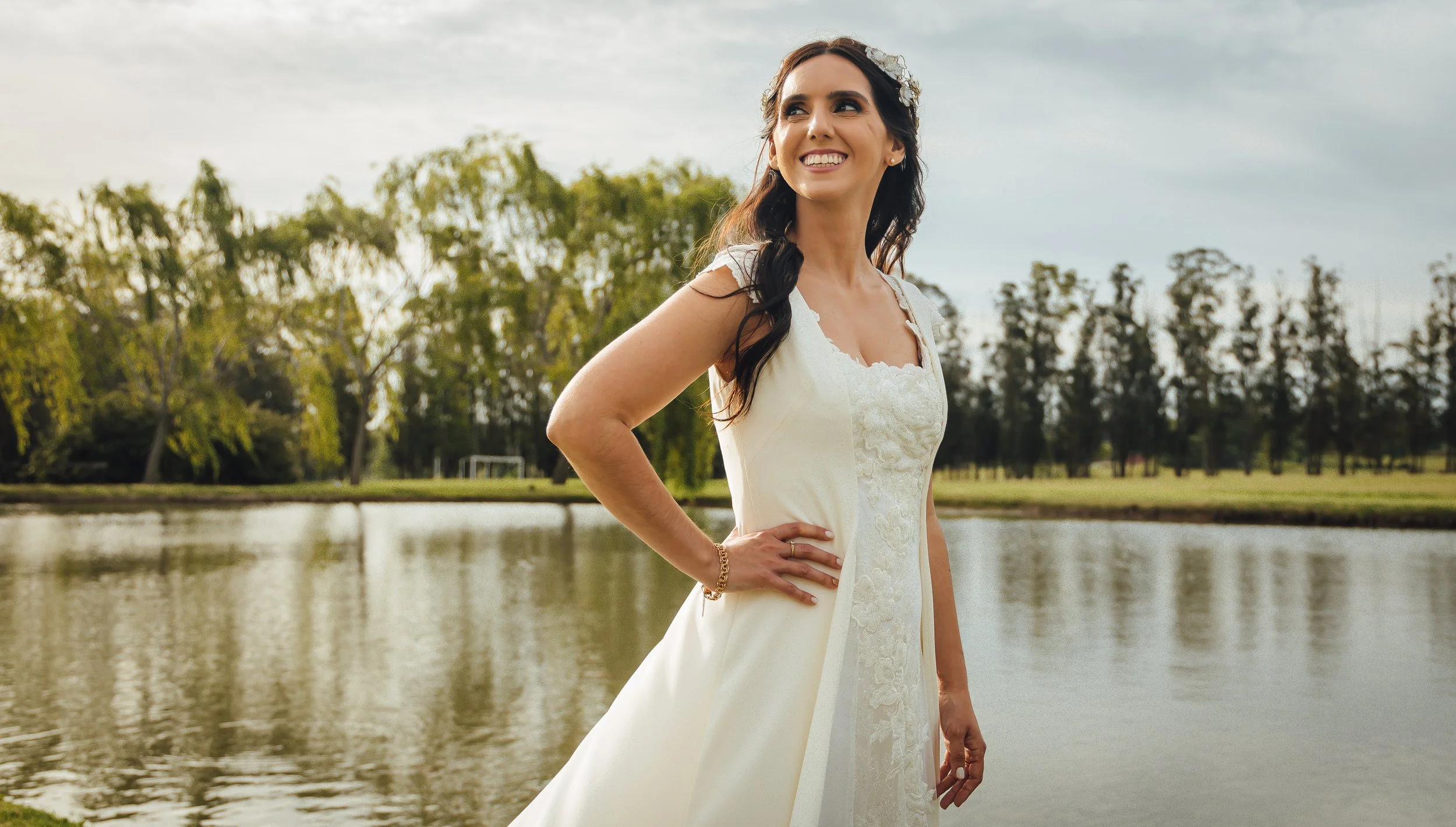 Mujer en vestido de boda blanco, sonriendo junto a un lago con árboles al fondo.