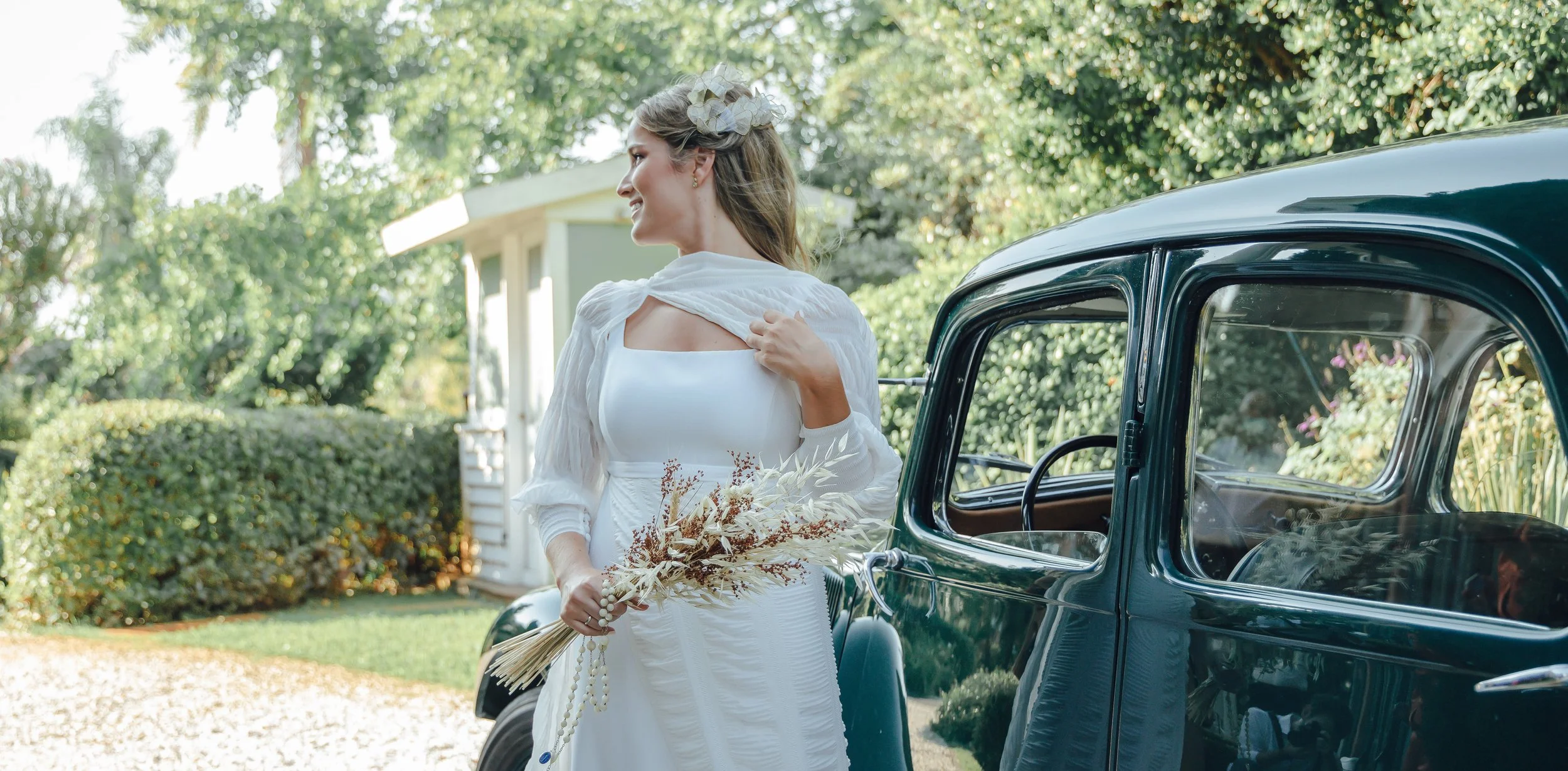 Novia en vestido blanco sosteniendo flores secas al lado de un auto clásico, con fondo de vegetación.