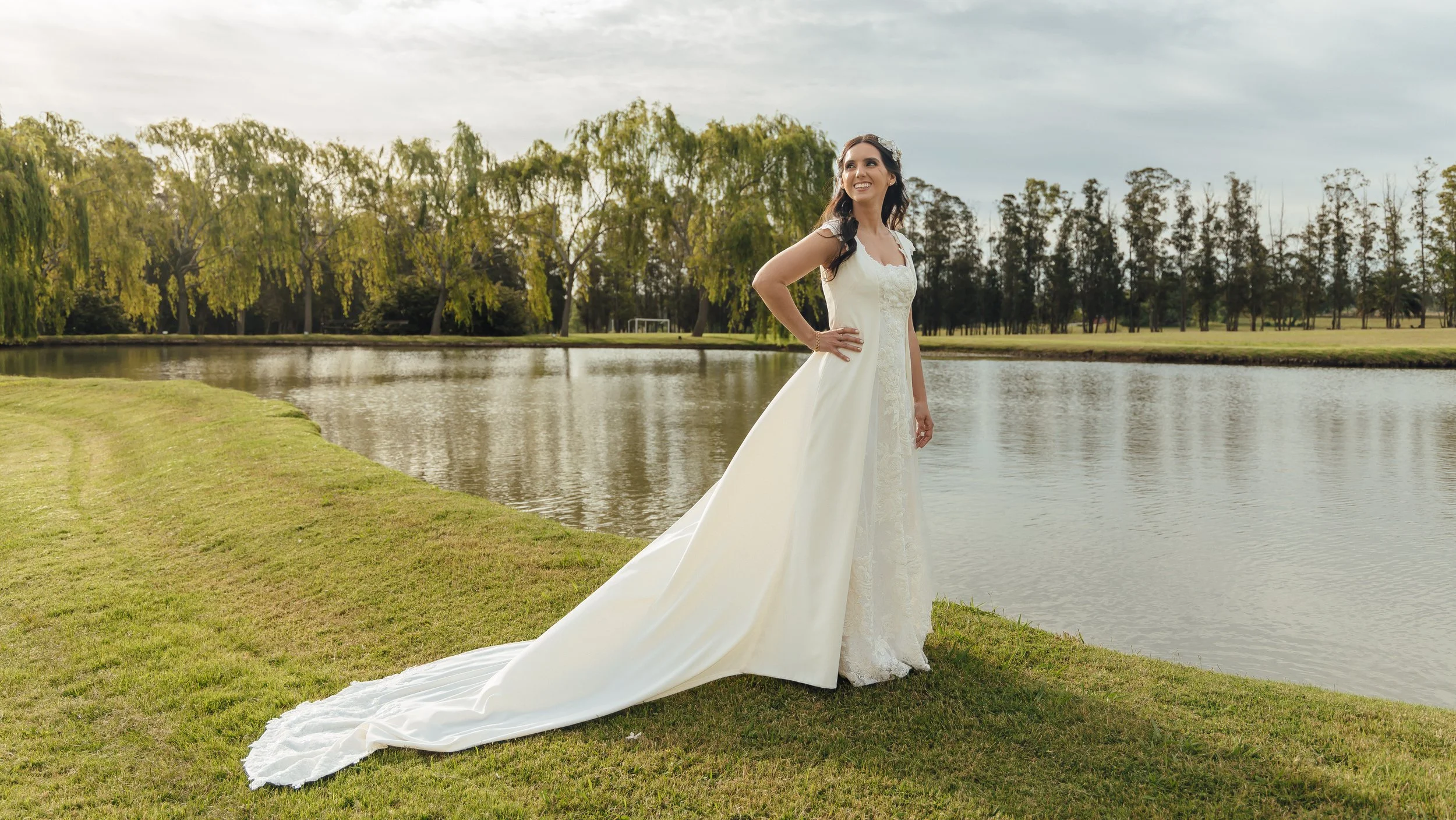 Novia con vestido blanco largo posando junto a un lago con árboles y césped alrededor, bajo un cielo nublado.
