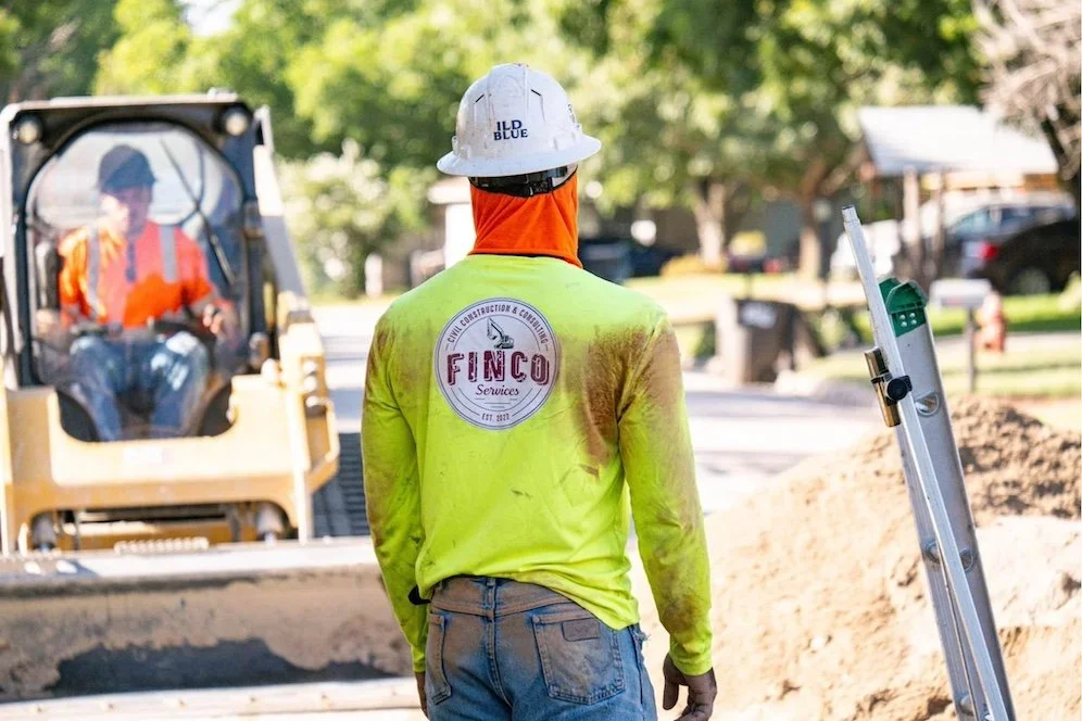 Construction worker wearing a yellow safety shirt with a logo on the back, a white hard hat, and jeans, standing on a road construction site with equipment and a worker in the background.