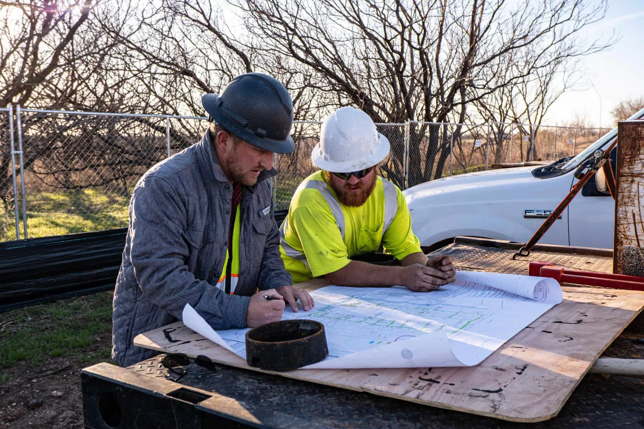 Two construction workers with helmets and safety vests looking at blueprints on a table outdoors, with a white pickup truck and leafless trees in the background.