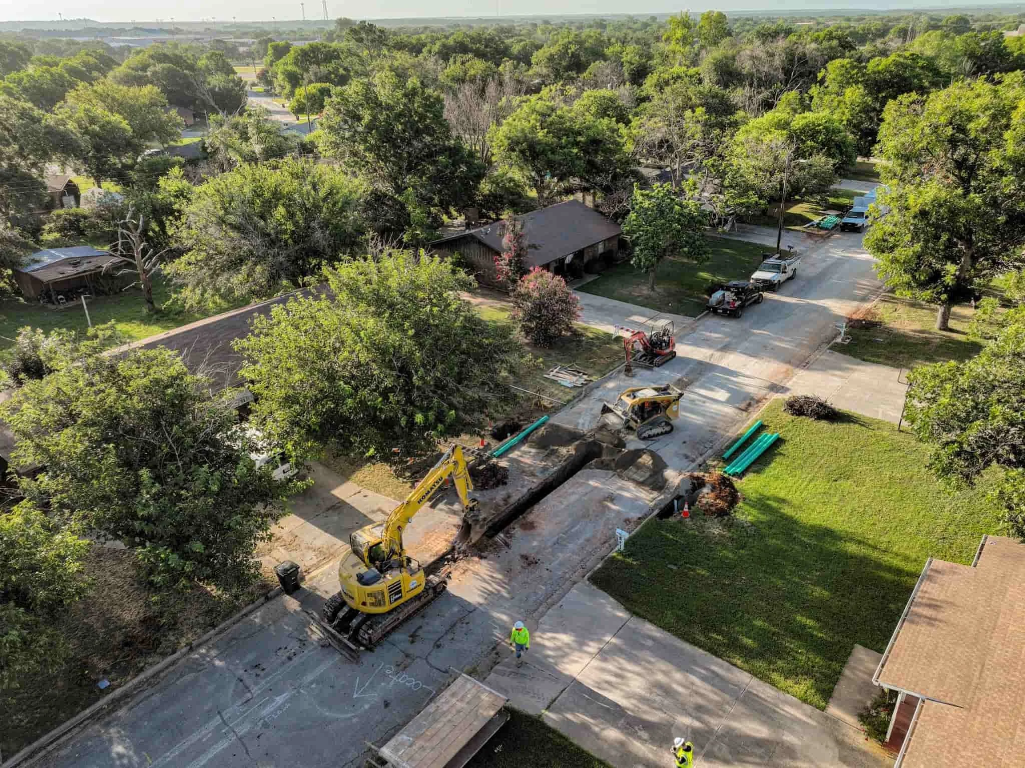 Construction workers repair a road with heavy machinery, including an excavator and compactors, in a suburban neighborhood with trees, houses, and parked trucks.