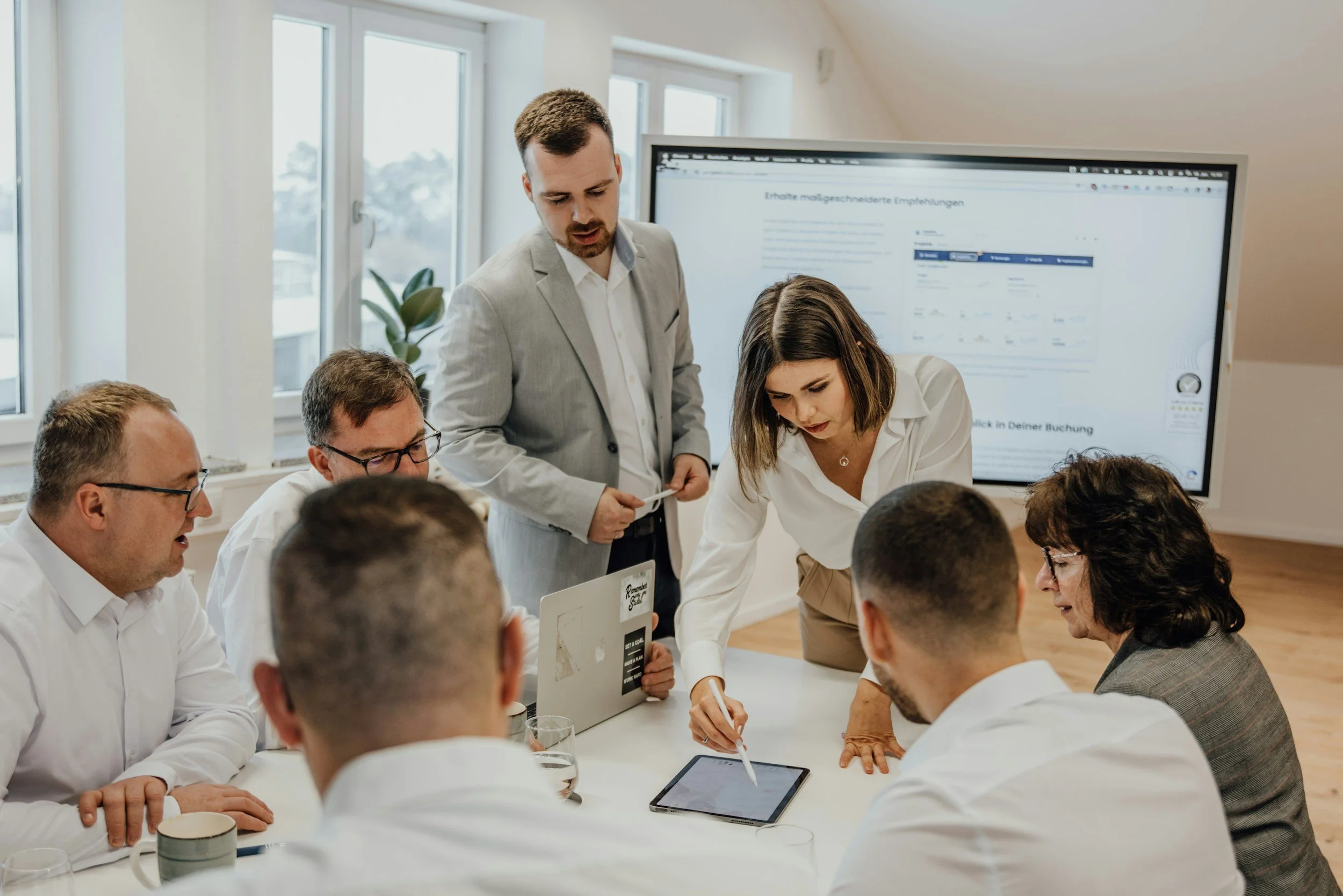 A group of seven business people gathered around a conference table in a meeting room, with a woman pointing at a tablet on the table and a man standing beside her, reviewing documents.