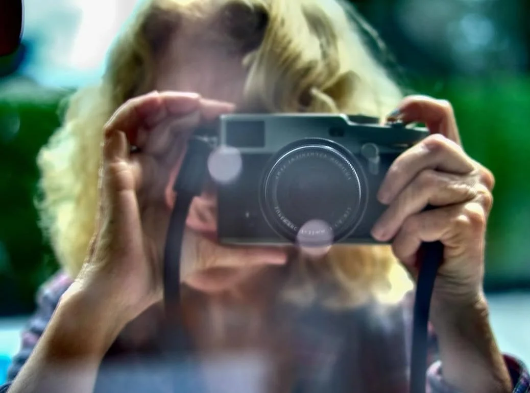 A person taking a photo with a camera reflected in a mirror. The person has blonde hair and is holding the camera close to their face.