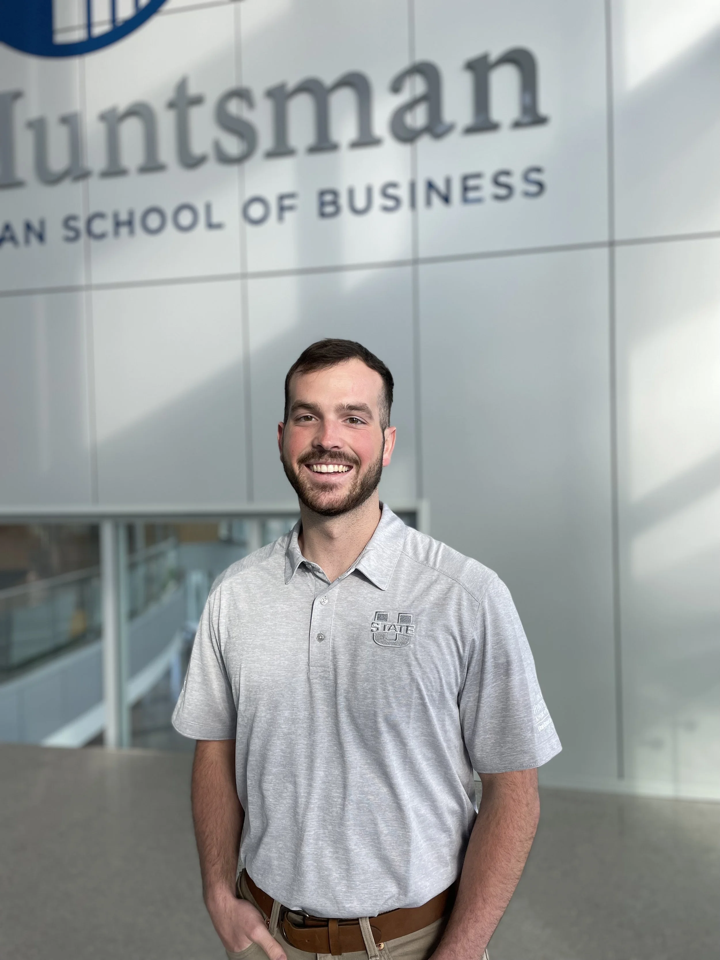A smiling young man in a gray polo shirt with a university logo standing in front of a large wall that reads 'Huntsman' and 'School of Business' at Utah State University.