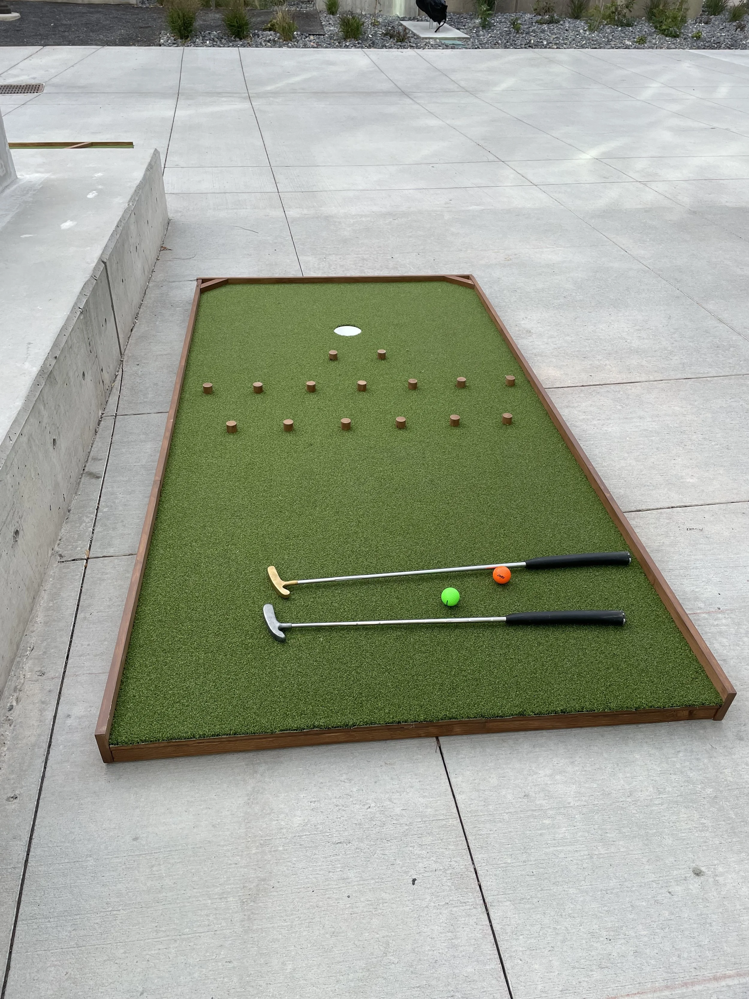 Mini golf putting green with two putters, a green ball, an orange ball, and several wooden balls, set on a concrete patio.