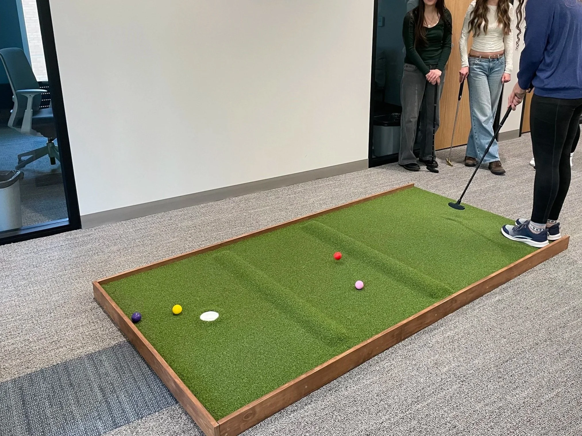 People playing indoor mini golf on a small green course in an office setting.