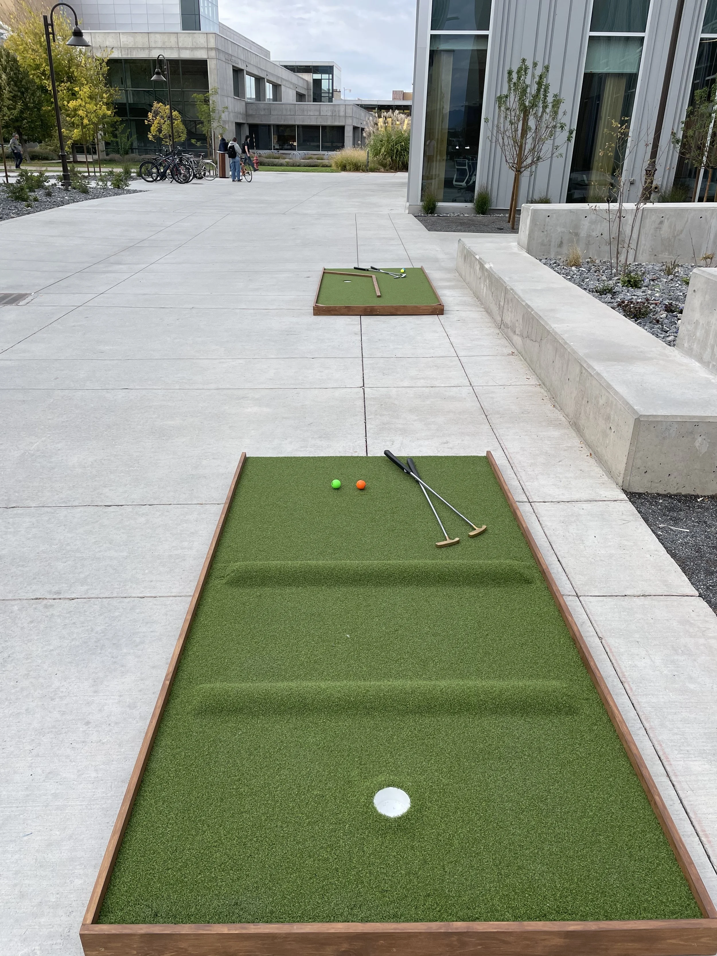 Outdoor mini golf course with two holes, featuring artificial turf and small golf clubs placed on the nearest hole. The course is set on a wide concrete walkway with modern buildings and trees in the background.