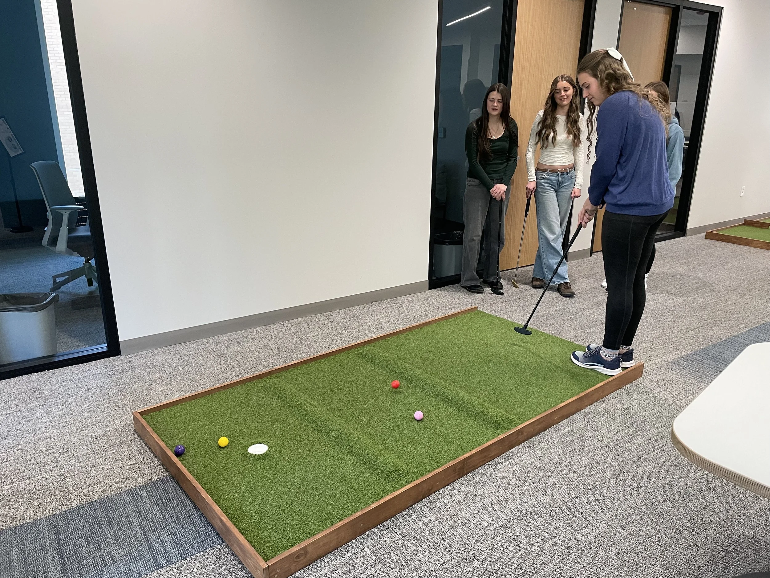 Group of young women playing mini golf indoors on a small green course.