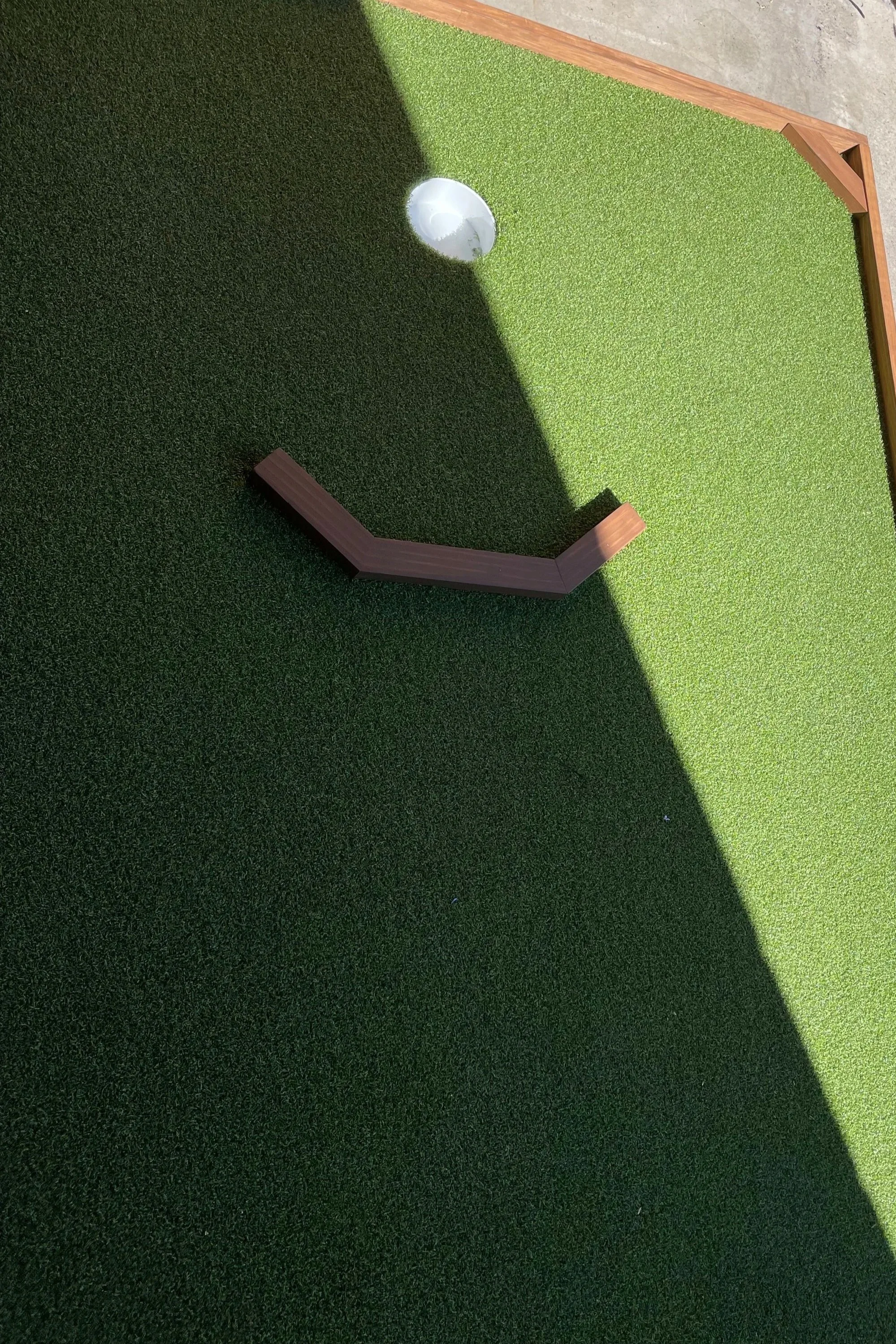 A mini golf course with a green artificial turf surface, a small wooden barrier, and a white plastic cup hole, partially shaded.