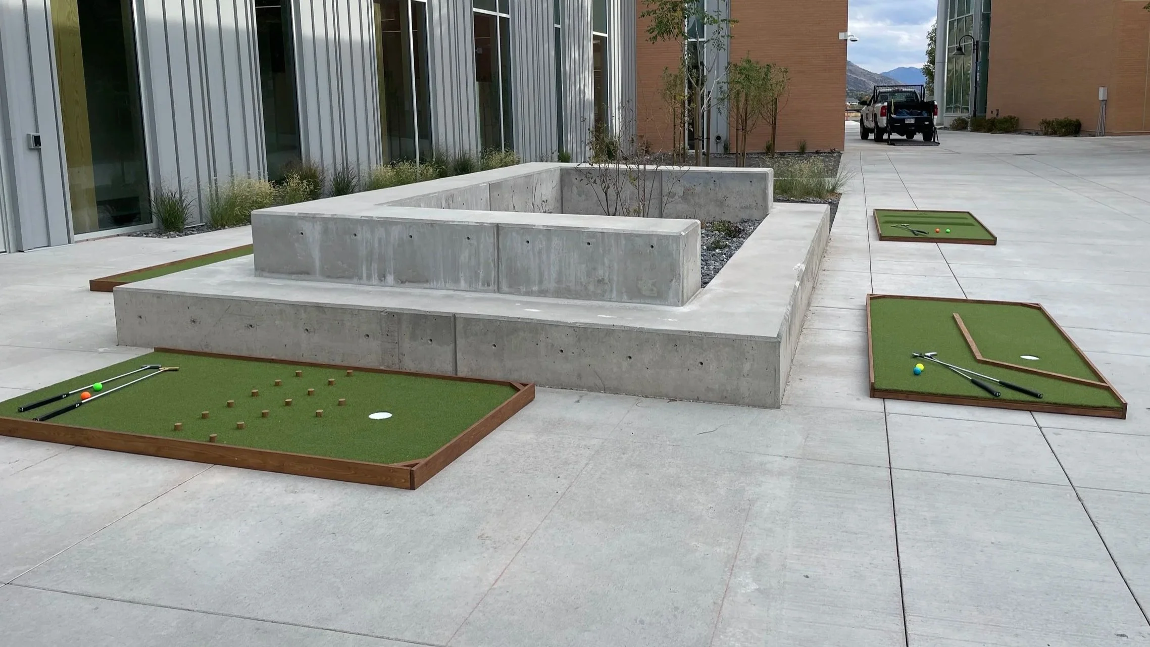 Outdoor mini golf putting greens with golf balls, putters, and obstacles, set on a paved area in front of modern buildings with concrete planters.